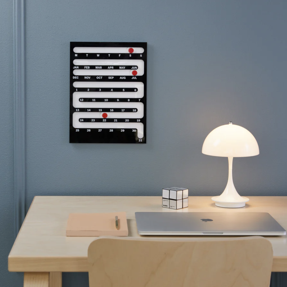  Perpetual Sliding Calendar featuring a maze-like path for a red dot to slide down, showing the current date. The calendar is on the wall next to a desk showing an Apple laptop, Rubik's cube, and white lamp. 