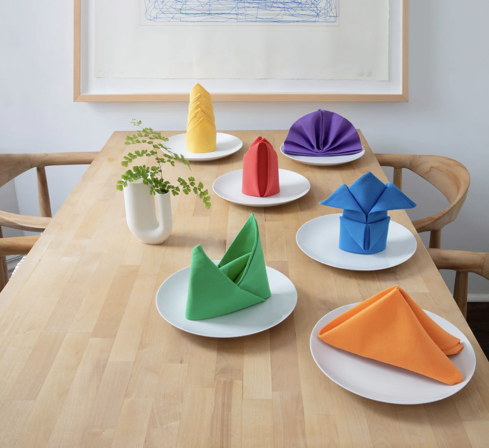  Wooden table with 6 white plates holding folded cloth napkins in yellow, orange, purple, blue, green and red. 