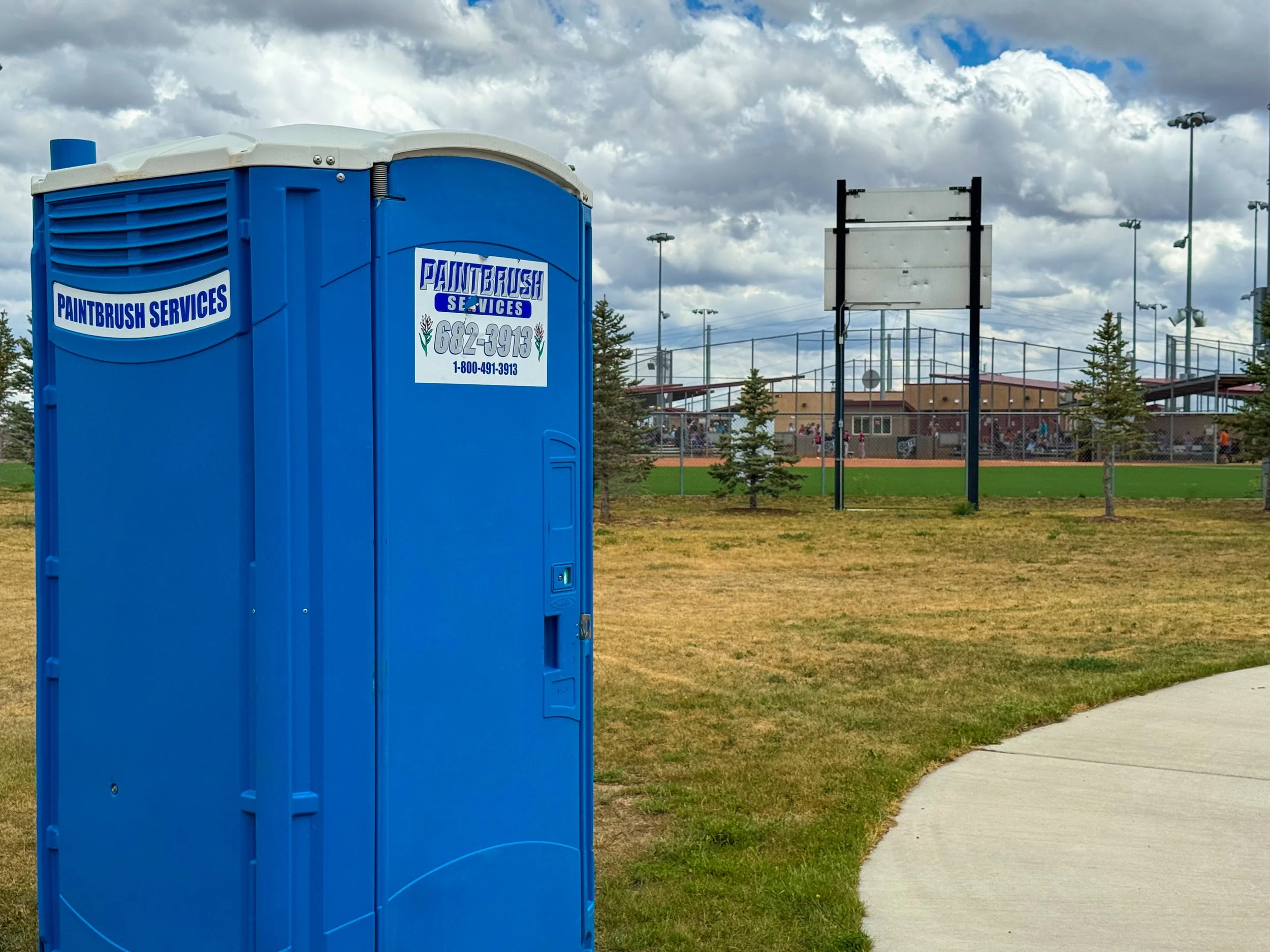 A blue portable toilet with a sign reading 'Paintbrush Services' in a park near a sports field with tall lights, a fence, and trees, under a cloudy sky.
