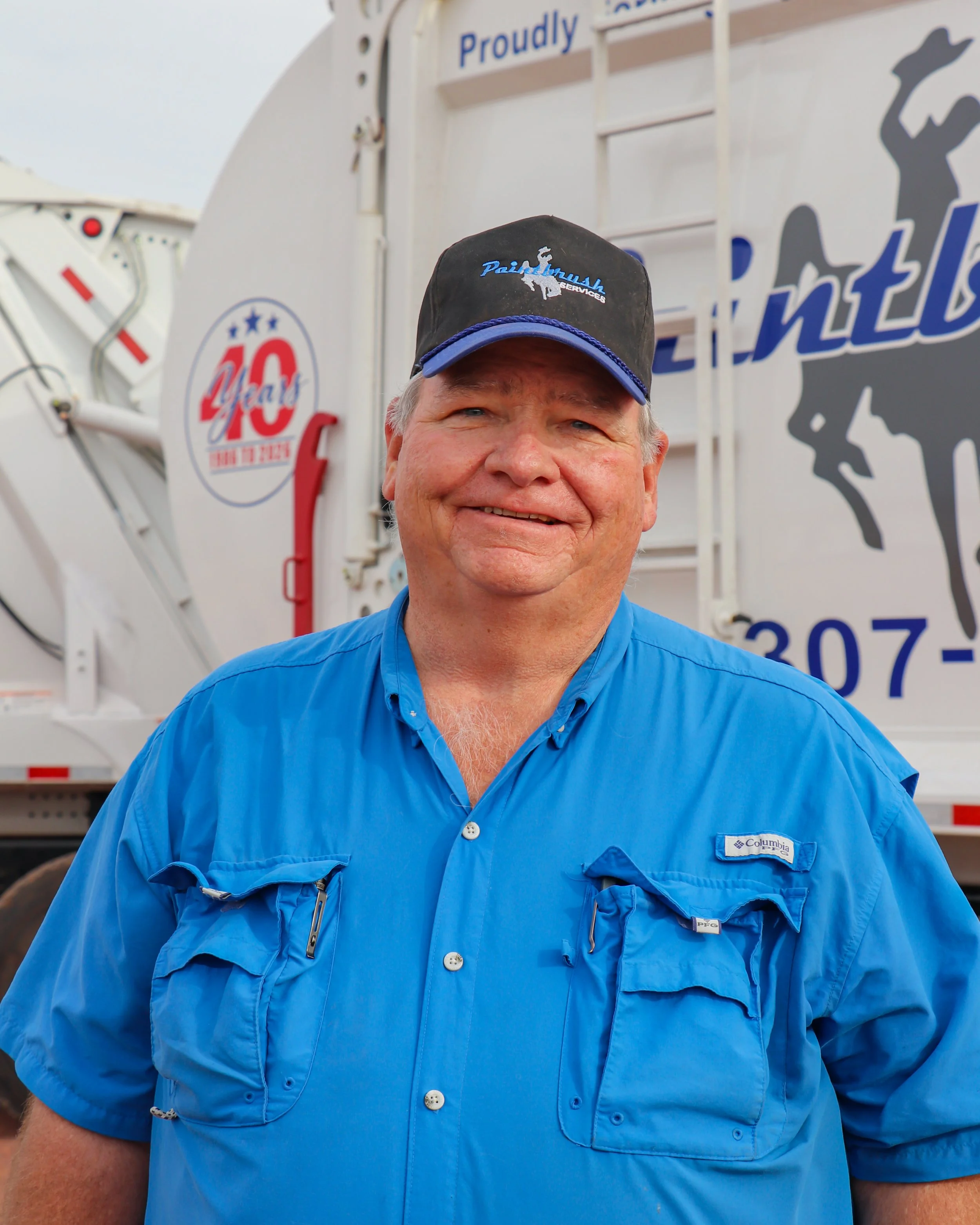 A smiling man wearing a black and blue Columbia jacket and a black baseball cap, standing in front of a large white delivery truck with logos and text, including the number 40 and company logo.
