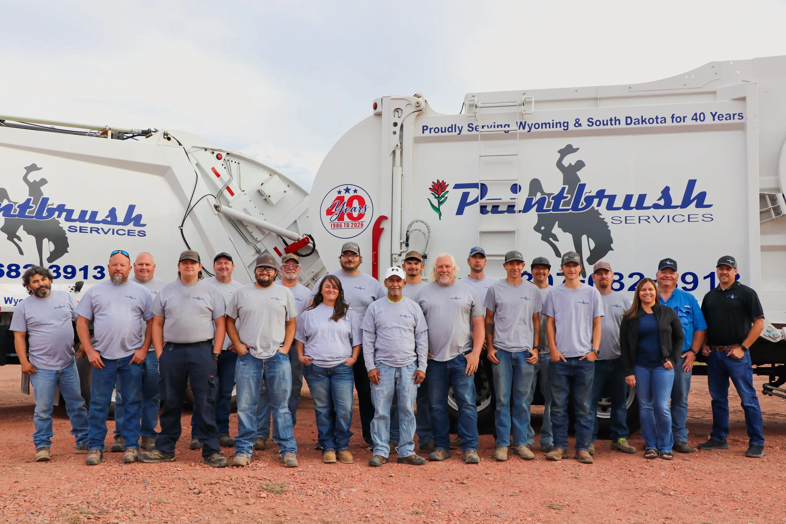 Group of people standing in front of a white waste collection truck with blue and red decals, some smiling and looking at the camera, on a red dirt ground with an overcast sky.
