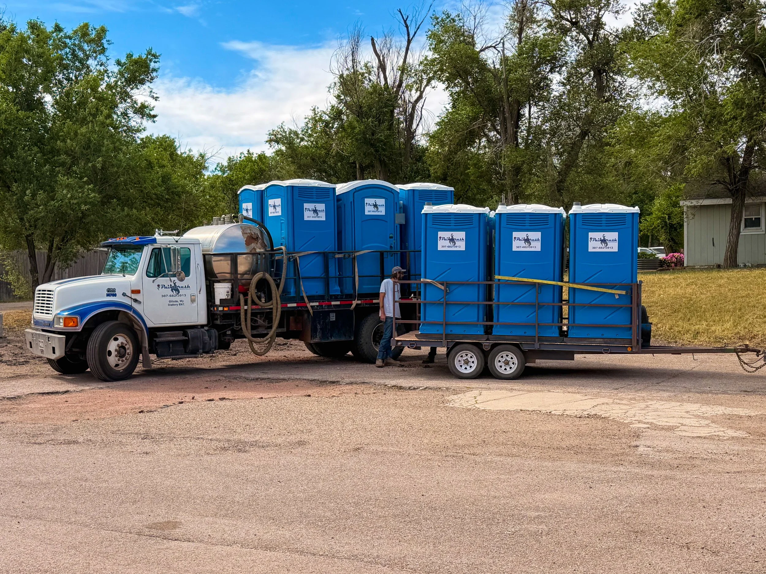 A white truck with blue portable toilets loaded on a trailer behind it, parked on a gravel road in a rural area with trees and a house in the background.