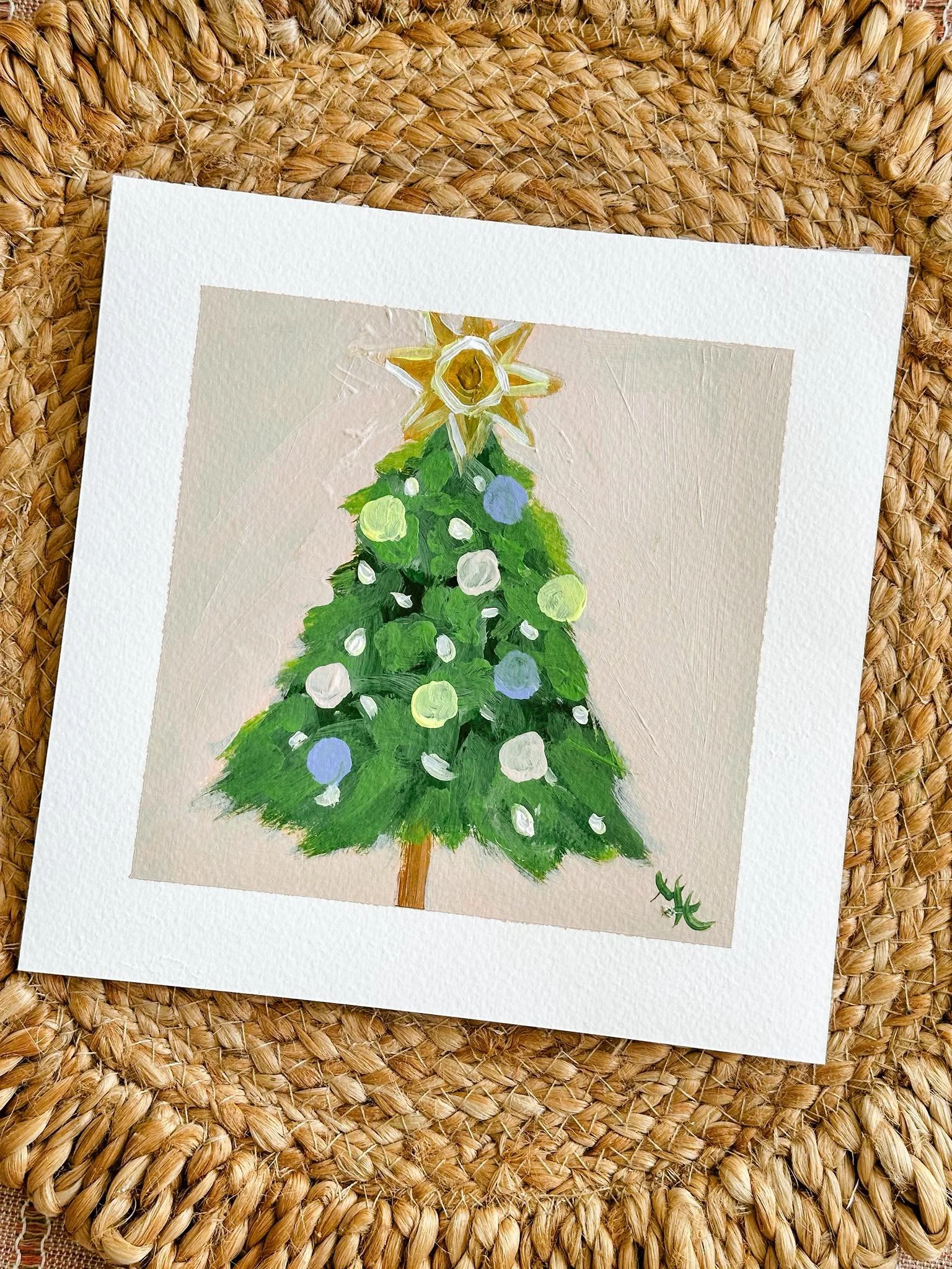 A hand-painted picture of a decorated Christmas tree with green branches, white and light blue ornaments, and a golden star on top, framed with a white border, placed on a woven straw surface.