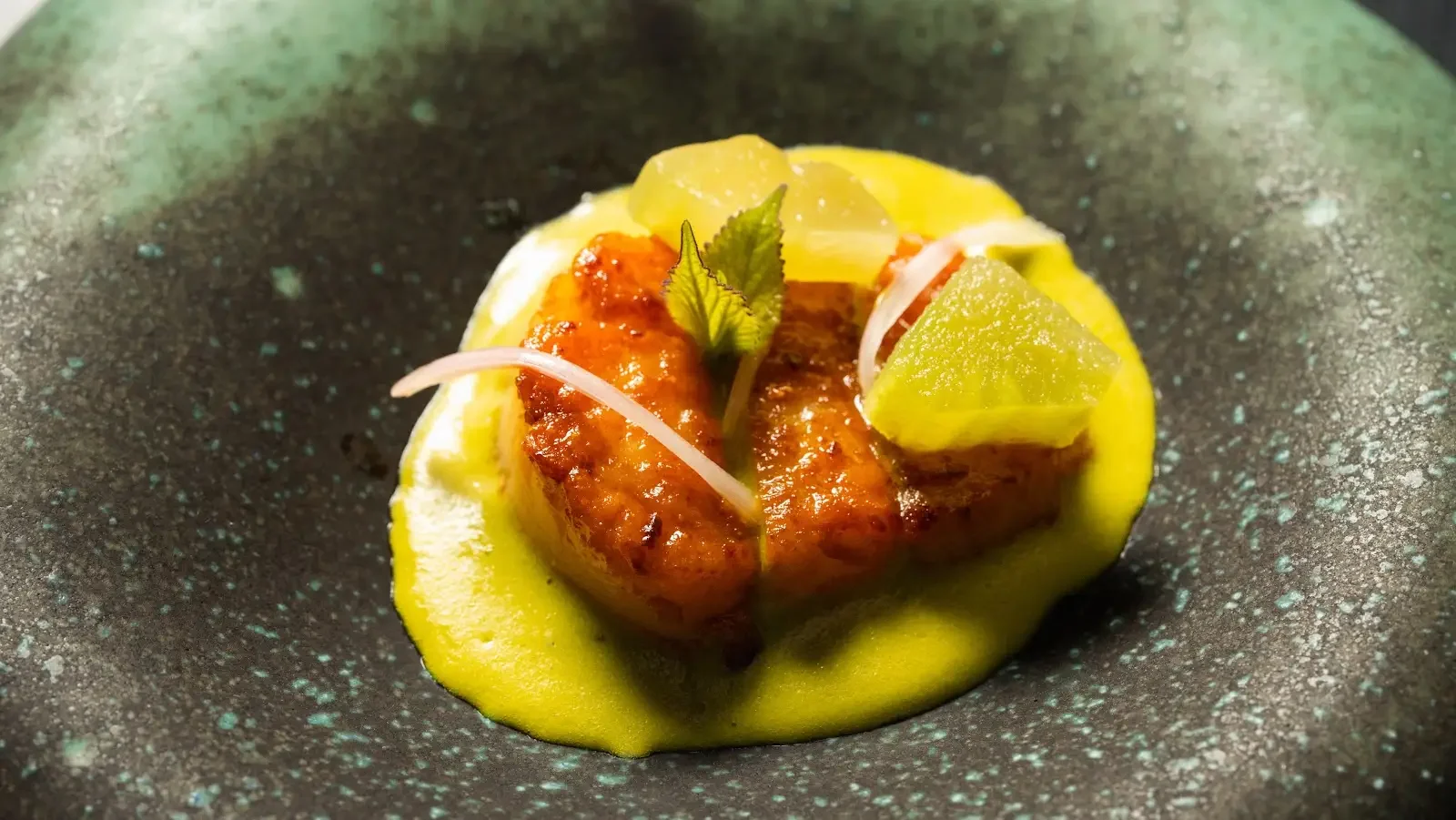 A gourmet dish with a piece of seared fish topped with sauce, garnished with herbs, served with a creamy yellow sauce and small pieces of fruit or vegetable on top, presented on a dark textured plate.