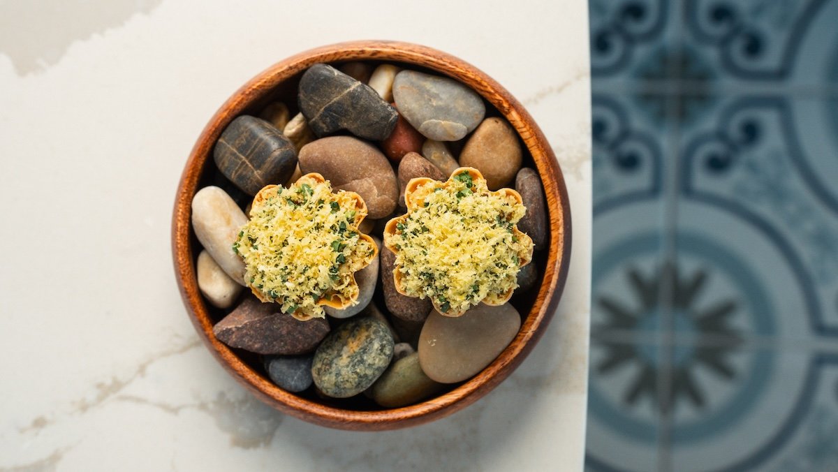 A round wooden bowl filled with various smooth, multicolored pebbles, with two flower-shaped small bites of food placed on top.
