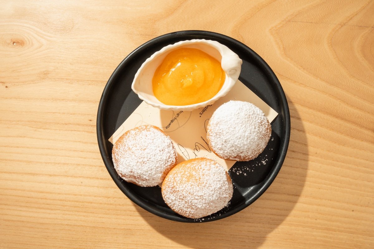 A black plate with three powdered sugar-coated croquettes and a small bowl of sauce on a wooden table.