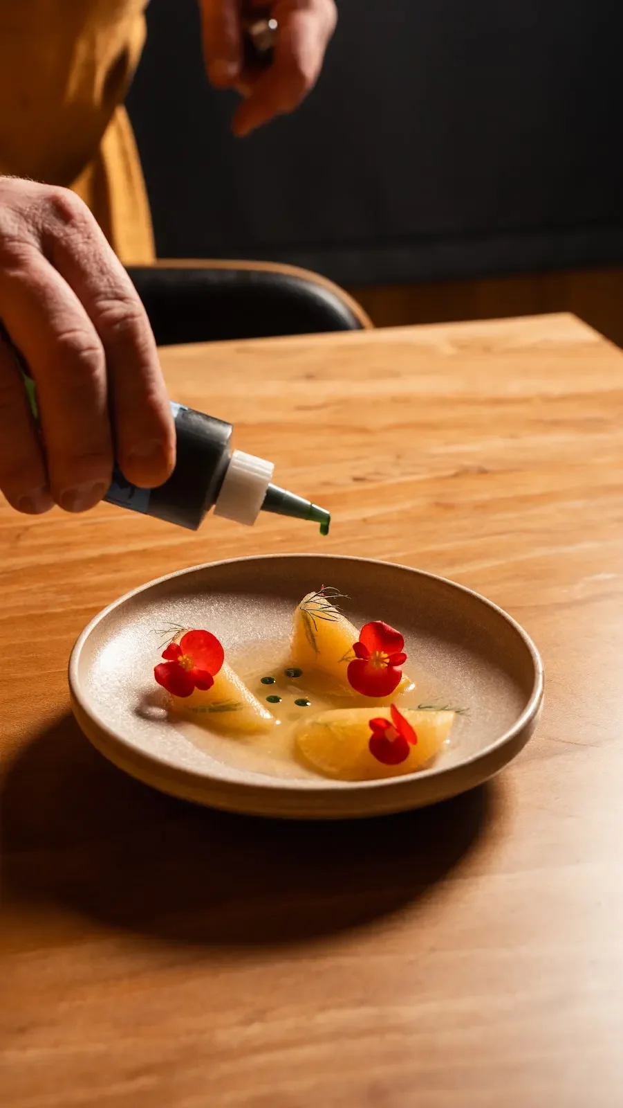 A chef pours a green sauce from a squeeze bottle onto a plated dish of sliced citrus fruit garnished with edible flowers and herbs on a small plate.