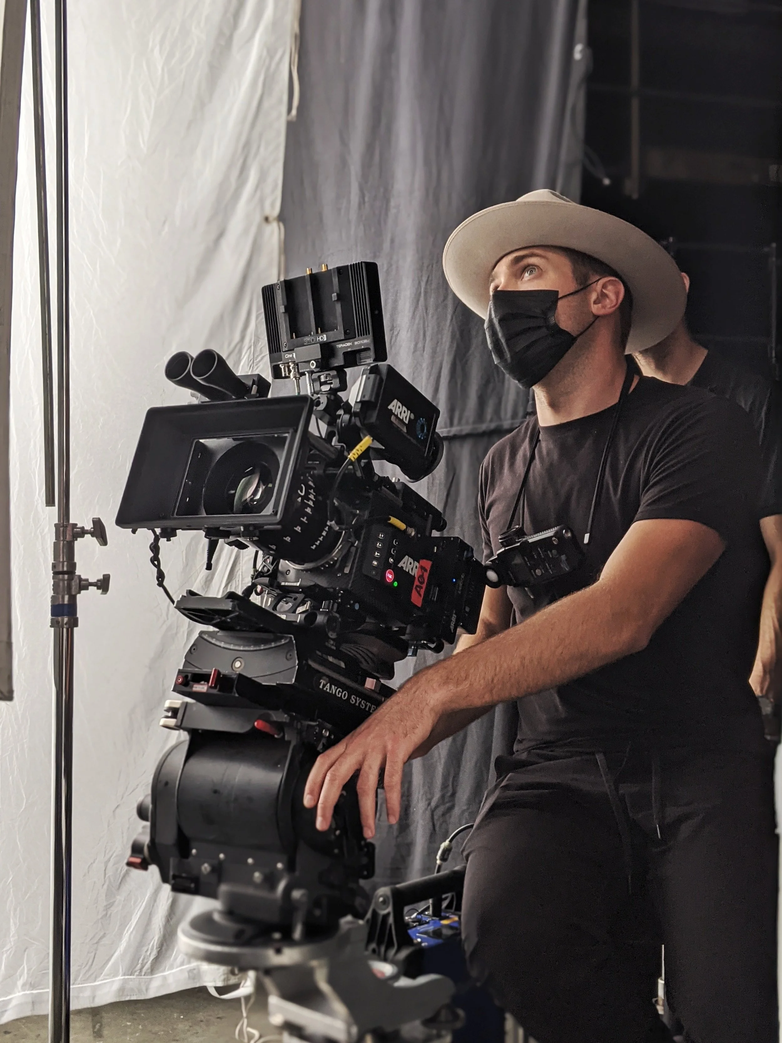 A man wearing a beige hat and a black face mask operates a professional video camera on a studio set with gray and white curtains.