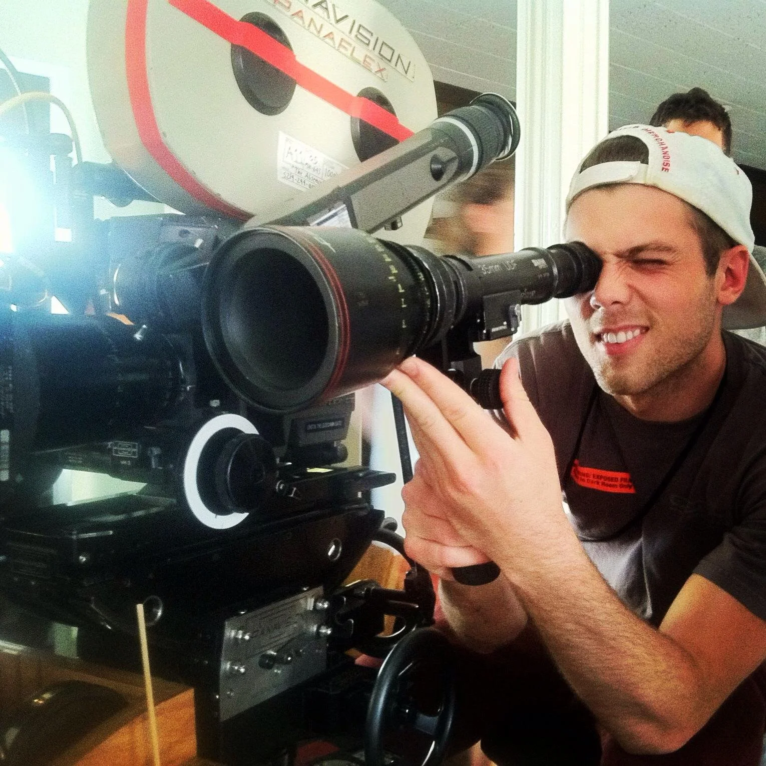 A man in a backward baseball cap looking through a professional camera's viewfinder, with a large reel of film or tape in the background.