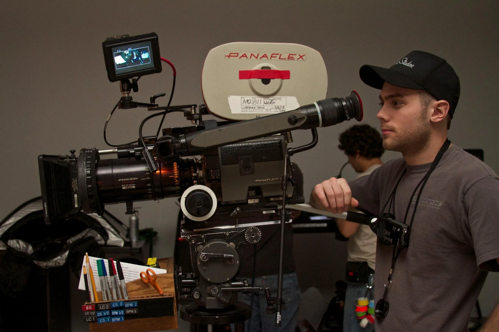 A person operating a professional film camera on a tripod, wearing a black cap and gray shirt, with a second person in the background. The camera has a large lens and an external monitor. Items such as pens, scissors, and color filters are on a nearb