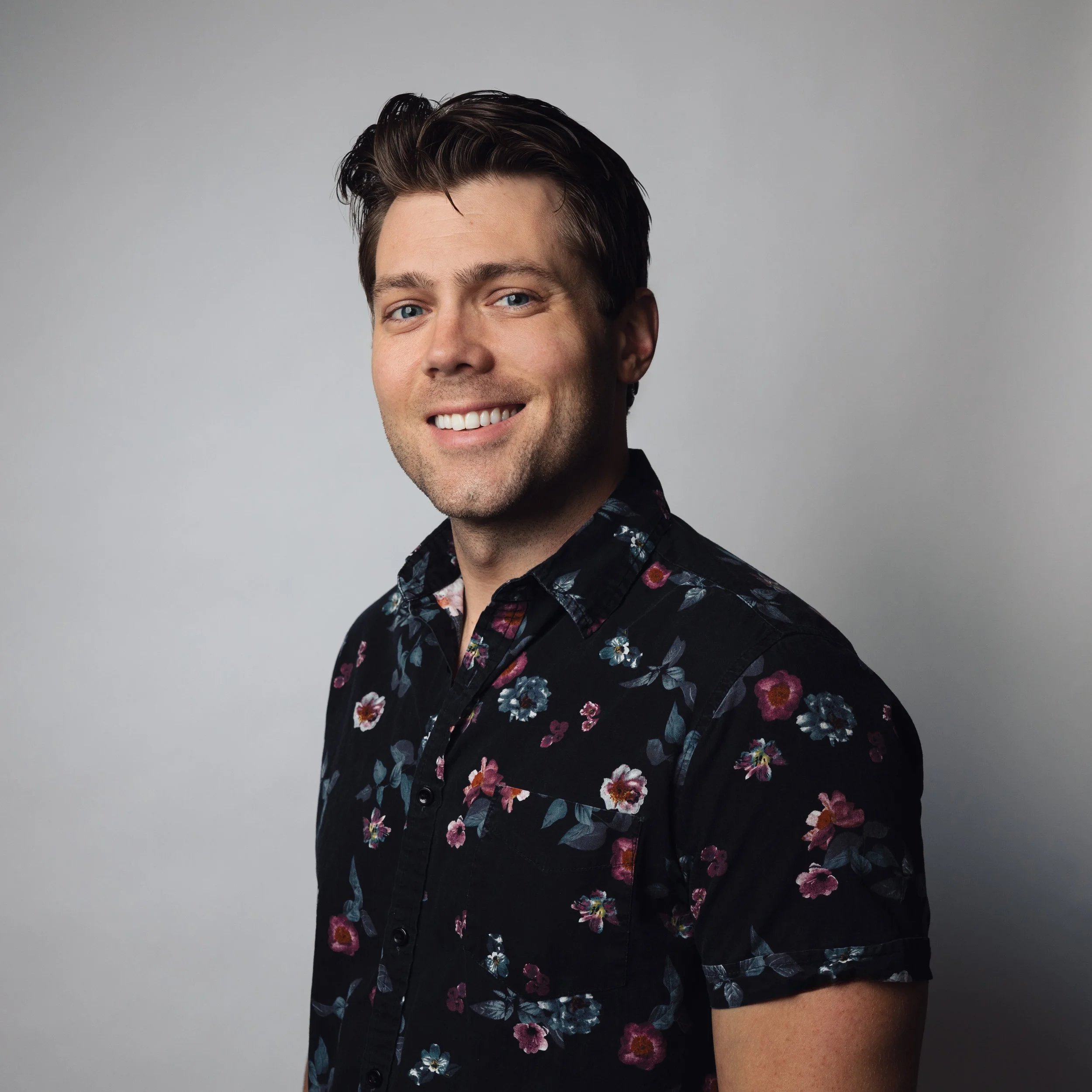 A smiling extremely handsome hunk of a man with dark hair and blue eyes wearing a black floral shirt against a plain gray background.