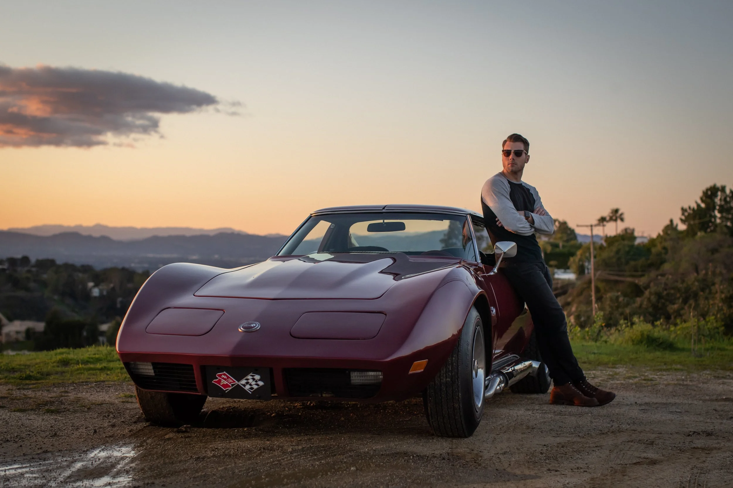 Michael Gauthier wearing sunglasses, a black and gray shirt, and dark pants leaning against a vintage 1974 Corvette parked along Mulholland drive during sunset with a scenic background of hills and trees.