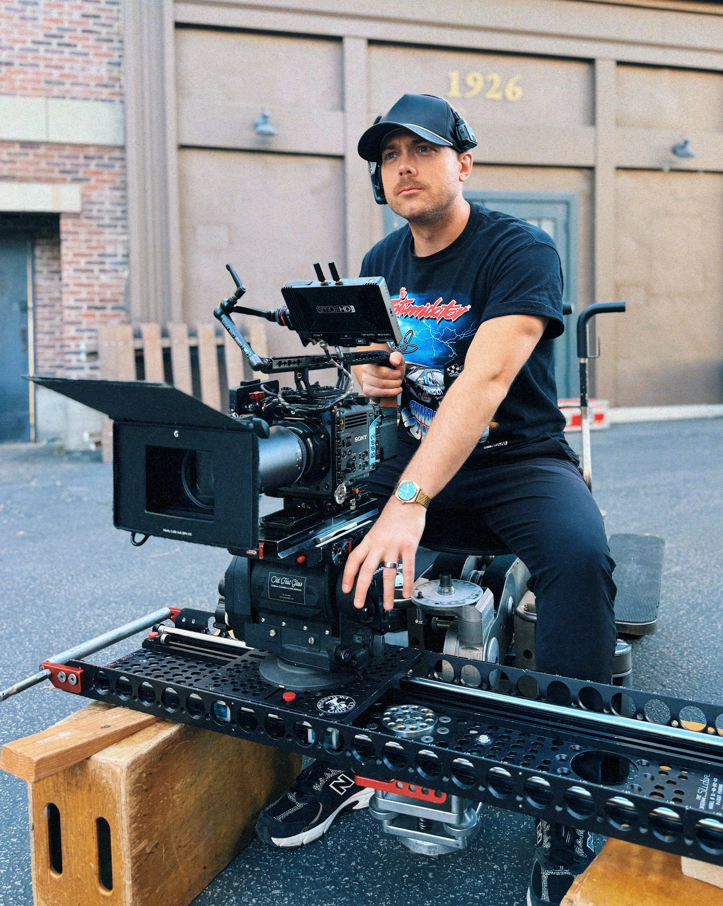 A man wearing a black cap, black t-shirt, and sneakers operating a professional camera mounted on a stabilizer rig outside in front of a building with the numbers 1926 visible. The camera is placed on a wooden support, and the man is adjusting the eq