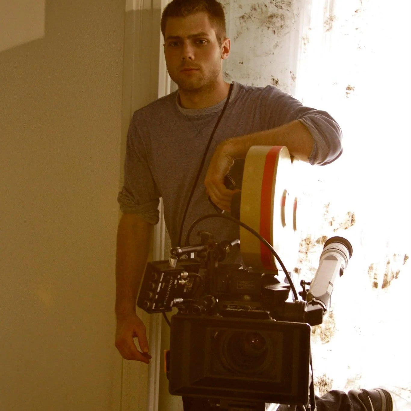 A man with short hair and a serious expression is taking a selfie in front of a mirror. He is holding what appears to be a camera or equipment. The background shows a bright window with curtains and some textured wall.