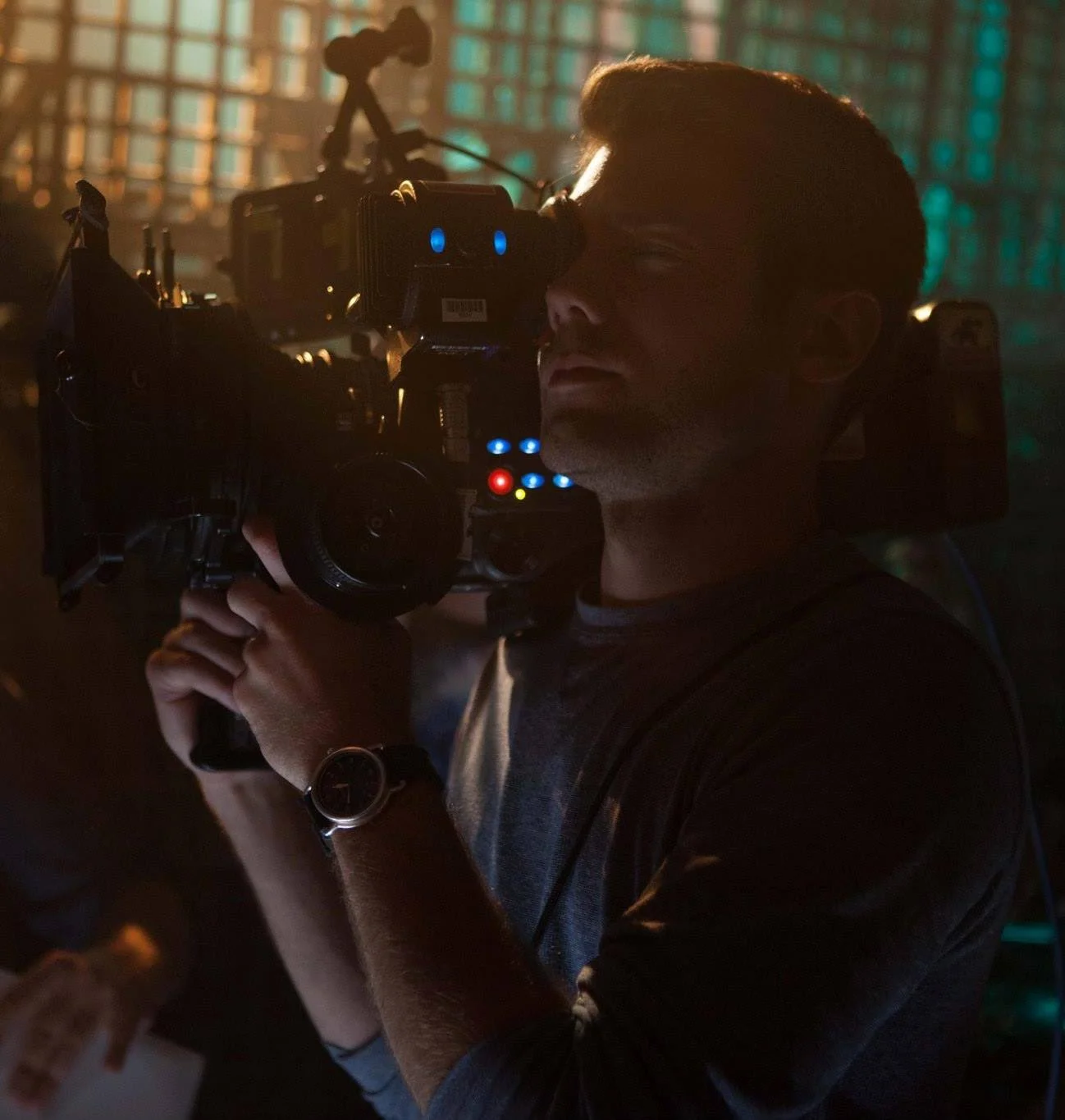 A person operating a professional film camera in a dimly lit setting with colorful, illuminated background.