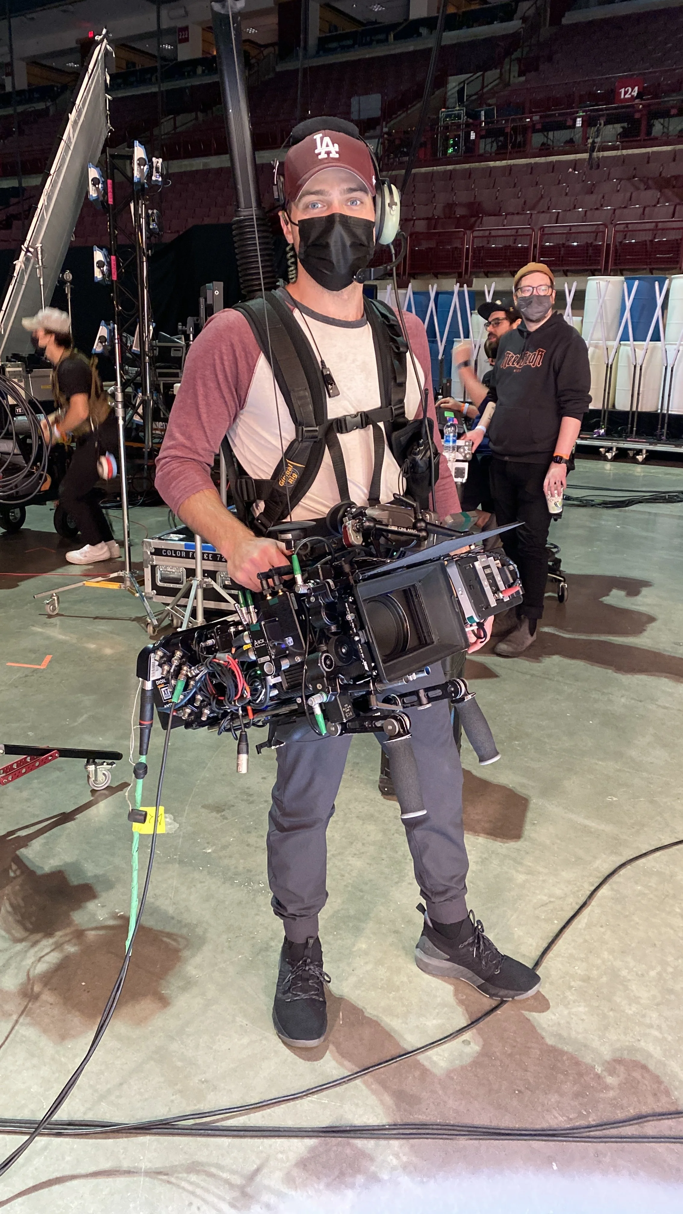 A man wearing a Los Angeles Dodgers cap and face mask holding a professional video camera inside an arena with empty red seats and crew members preparing equipment.