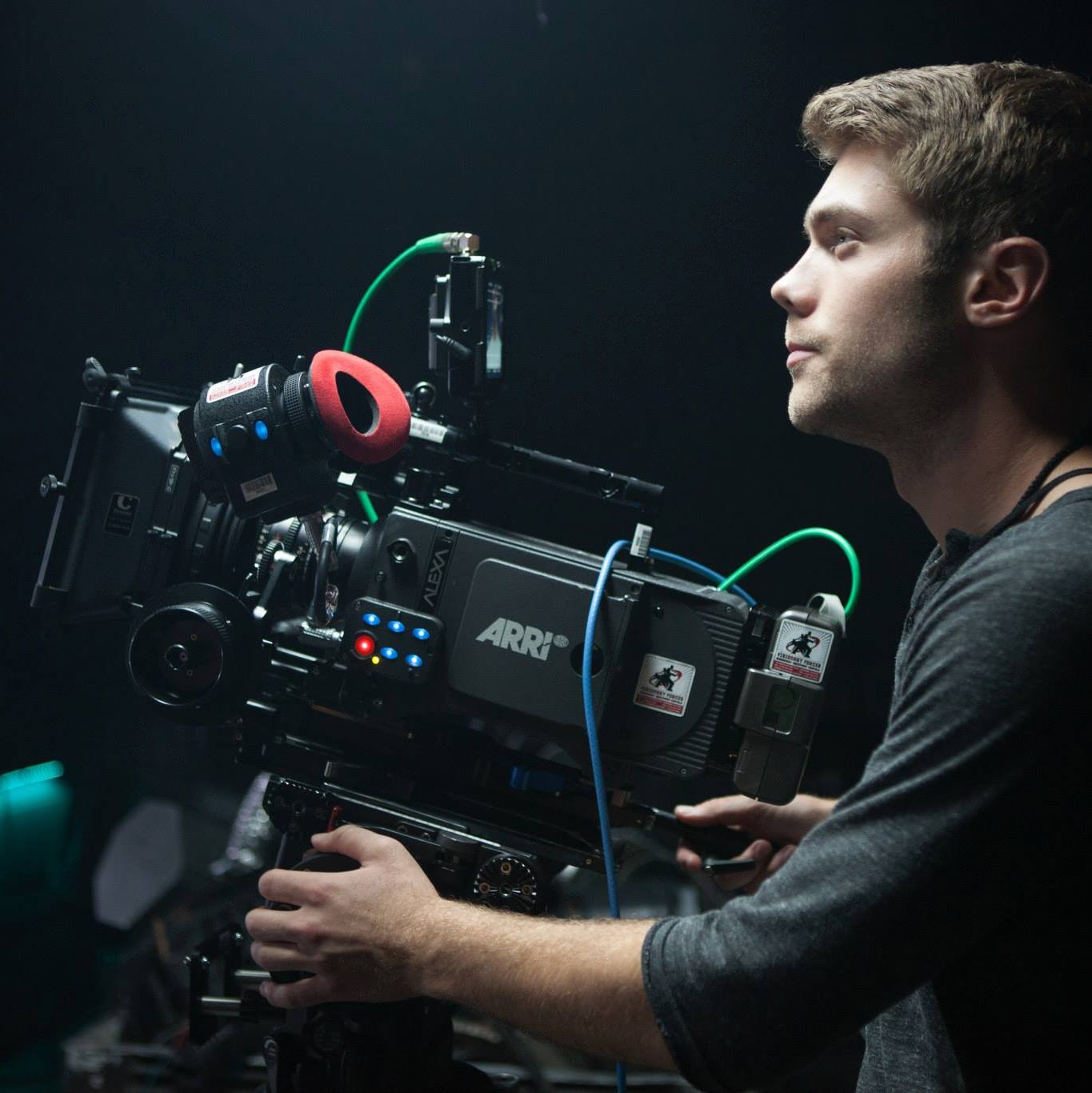 A man holds a professional film camera on a film set, with a dark background.