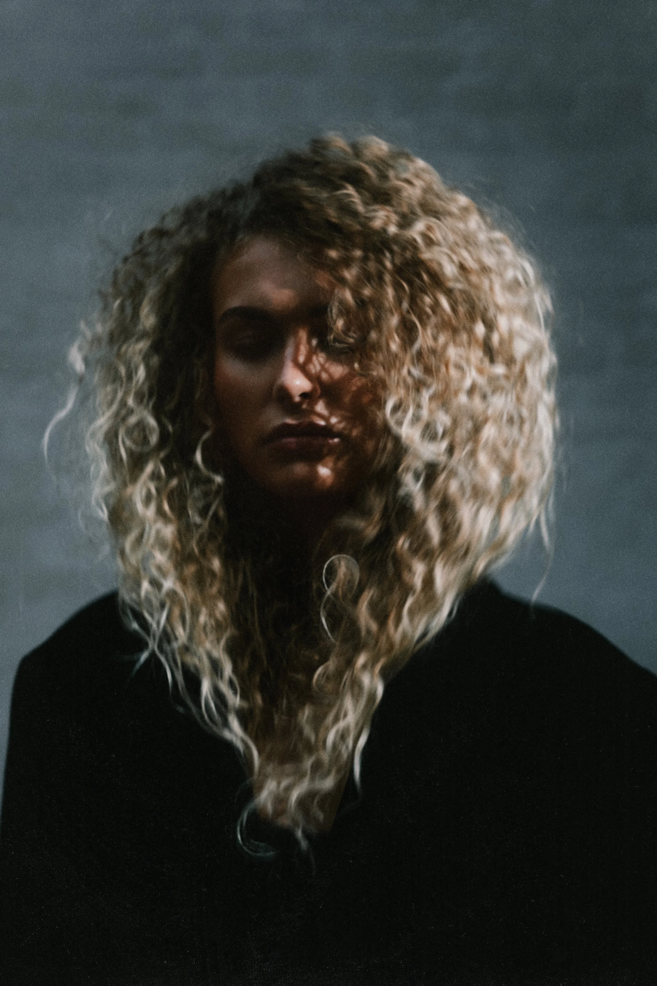 A woman with long, curly blonde hair, wearing a black top, against a dark background.
