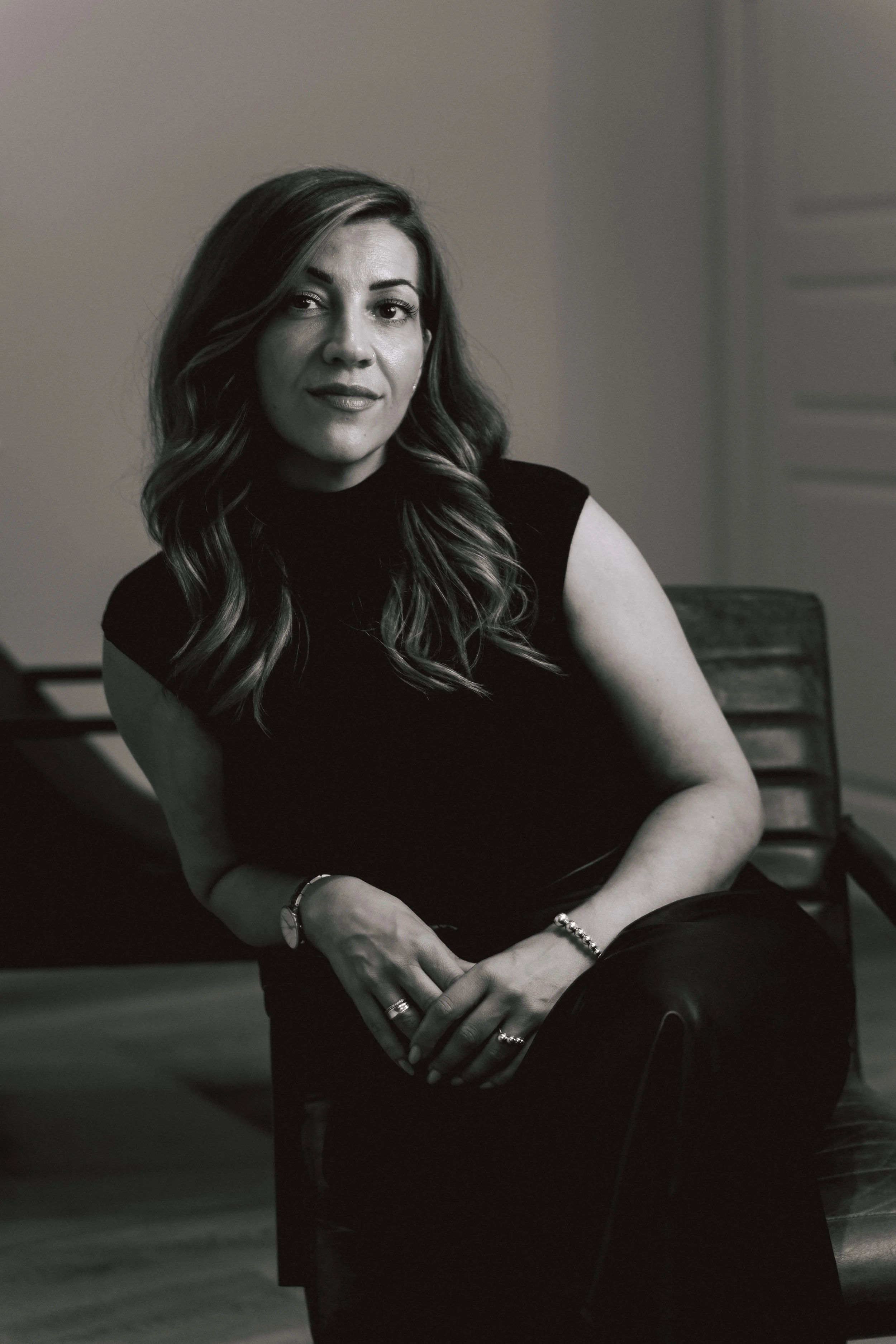 Black and white portrait of a woman with wavy hair, wearing a sleeveless top, jewelry including rings, bracelet, and watch, sitting on a chair with a blank wall and part of a window in the background.