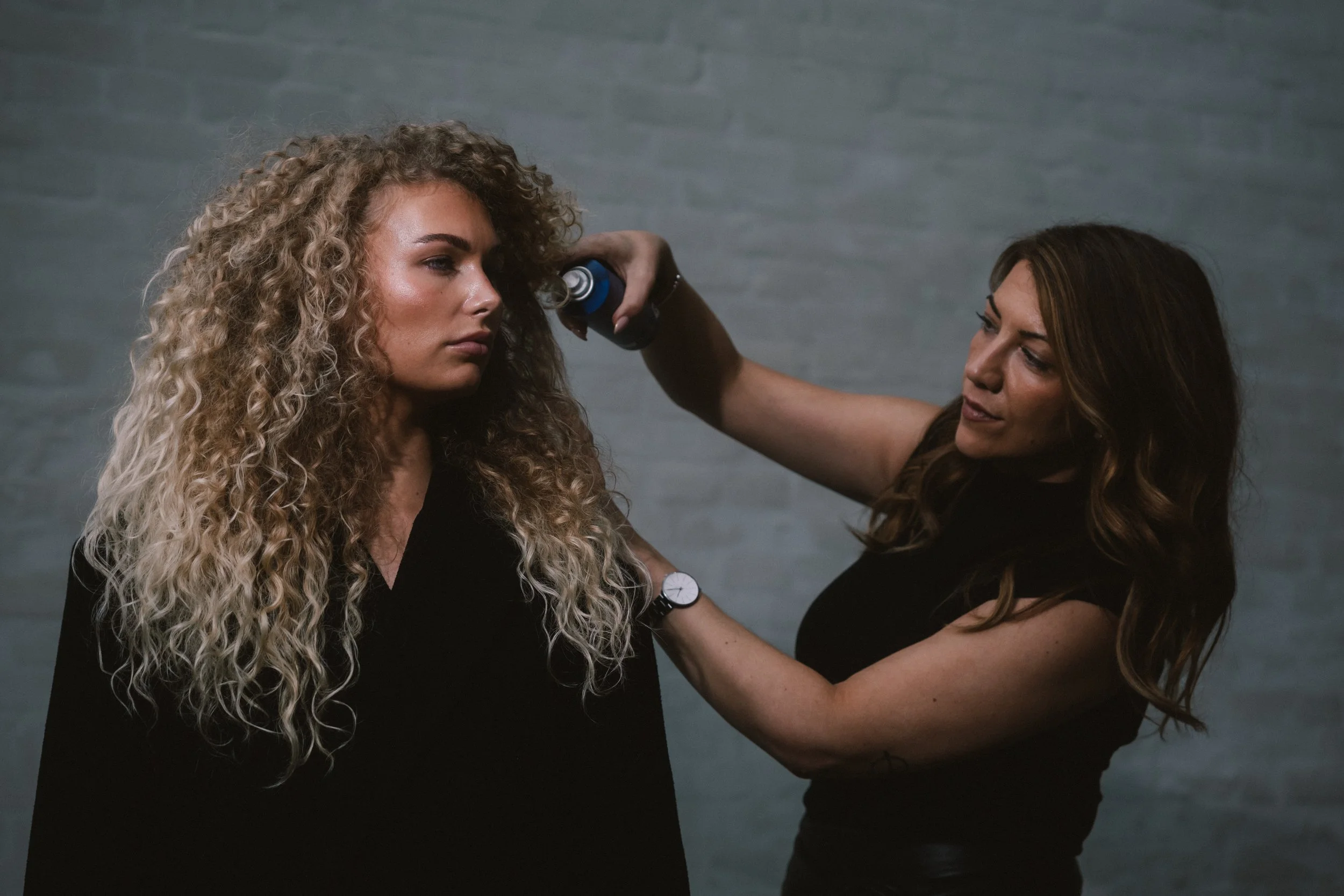 A woman with curly blonde hair has her face examined by a professional with brown hair, glasses, and a watch, against a gray brick wall background.