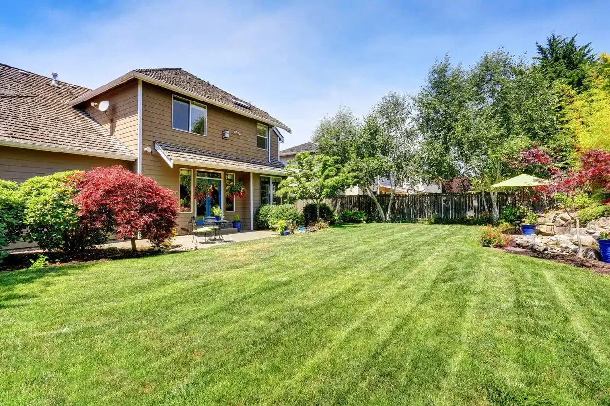 Spacious backyard with well-maintained green lawn, trees, shrubs, and a two-story house with beige siding, multiple windows, and a patio area with outdoor furniture and potted plants.
