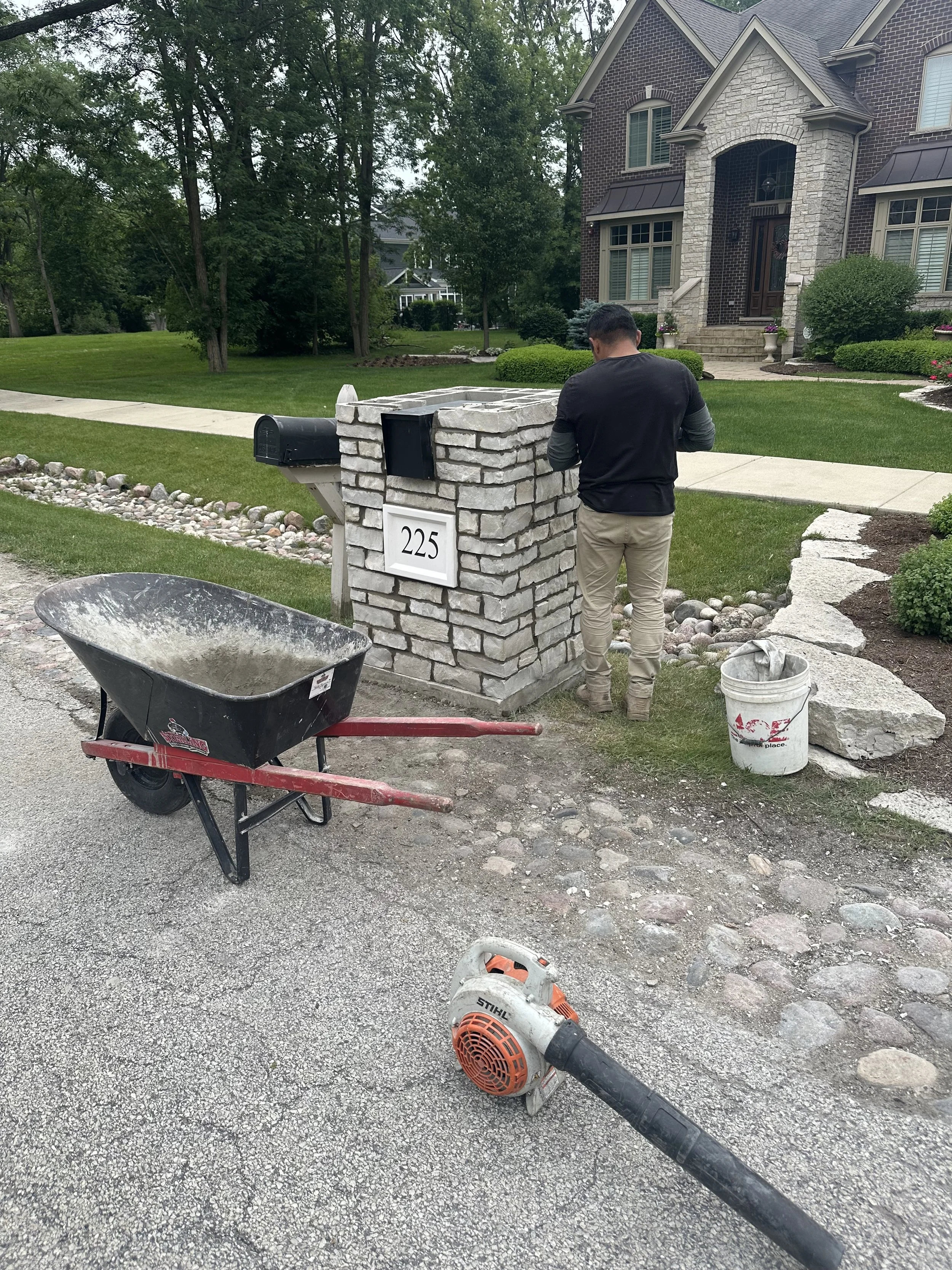 Person installing or repairing a brick mailbox at a residential home with equipment and tools nearby, including a wheelbarrow, leaf blower, and bucket.