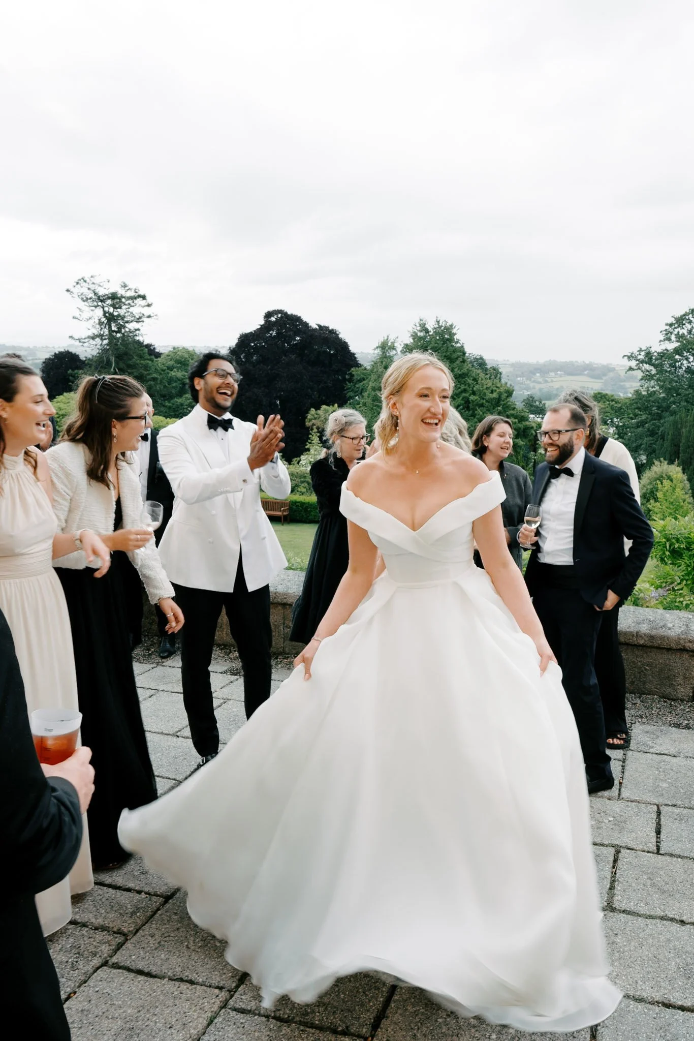 Bride in a white wedding dress smiling, surrounded by guests in formal attire outdoors on a cloudy day with greenery in the background.
