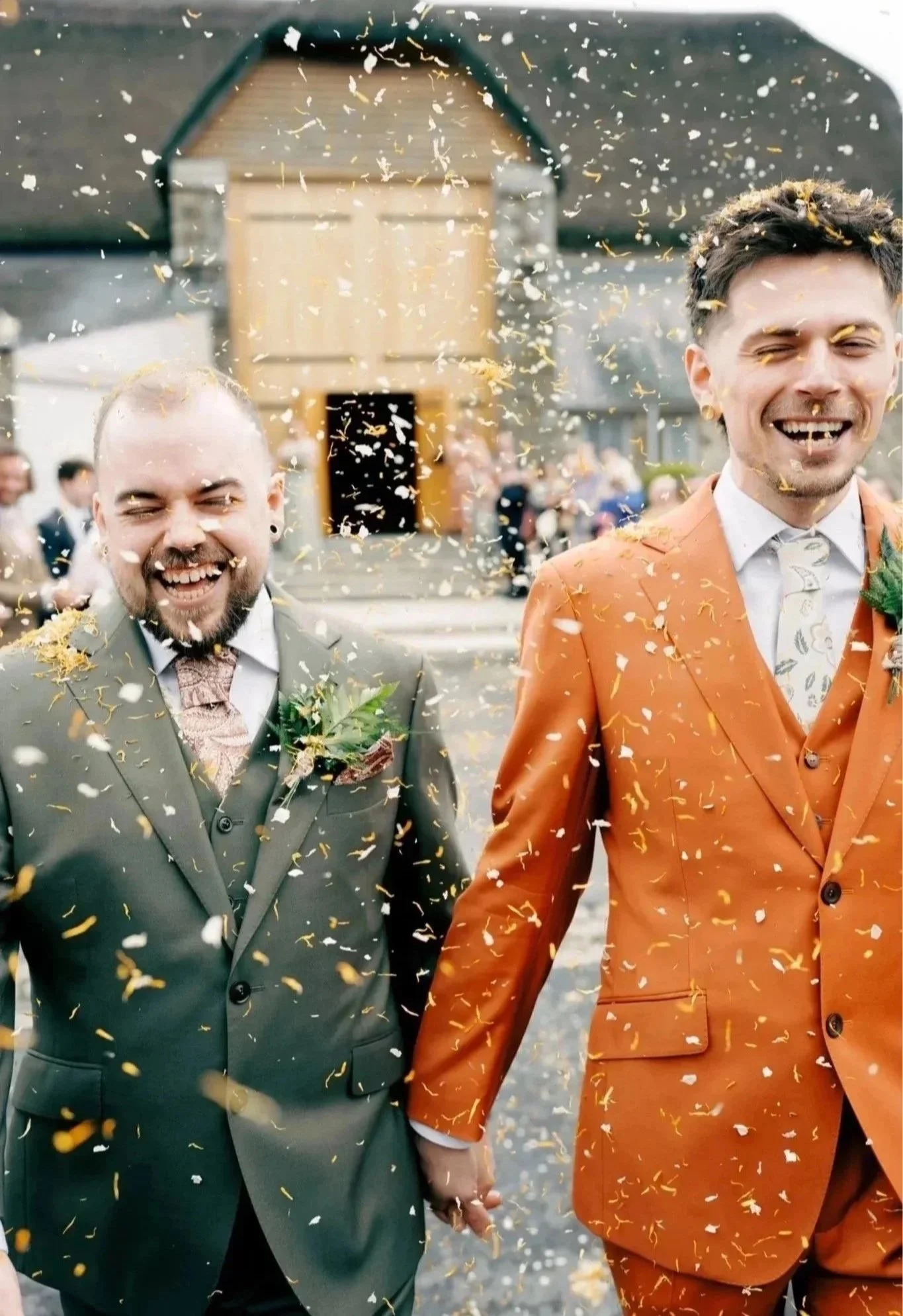 Two men dressed in suits, one in a gray suit and the other in an orange suit, holding hands and smiling as they walk through a shower of confetti at an outdoor wedding celebration.