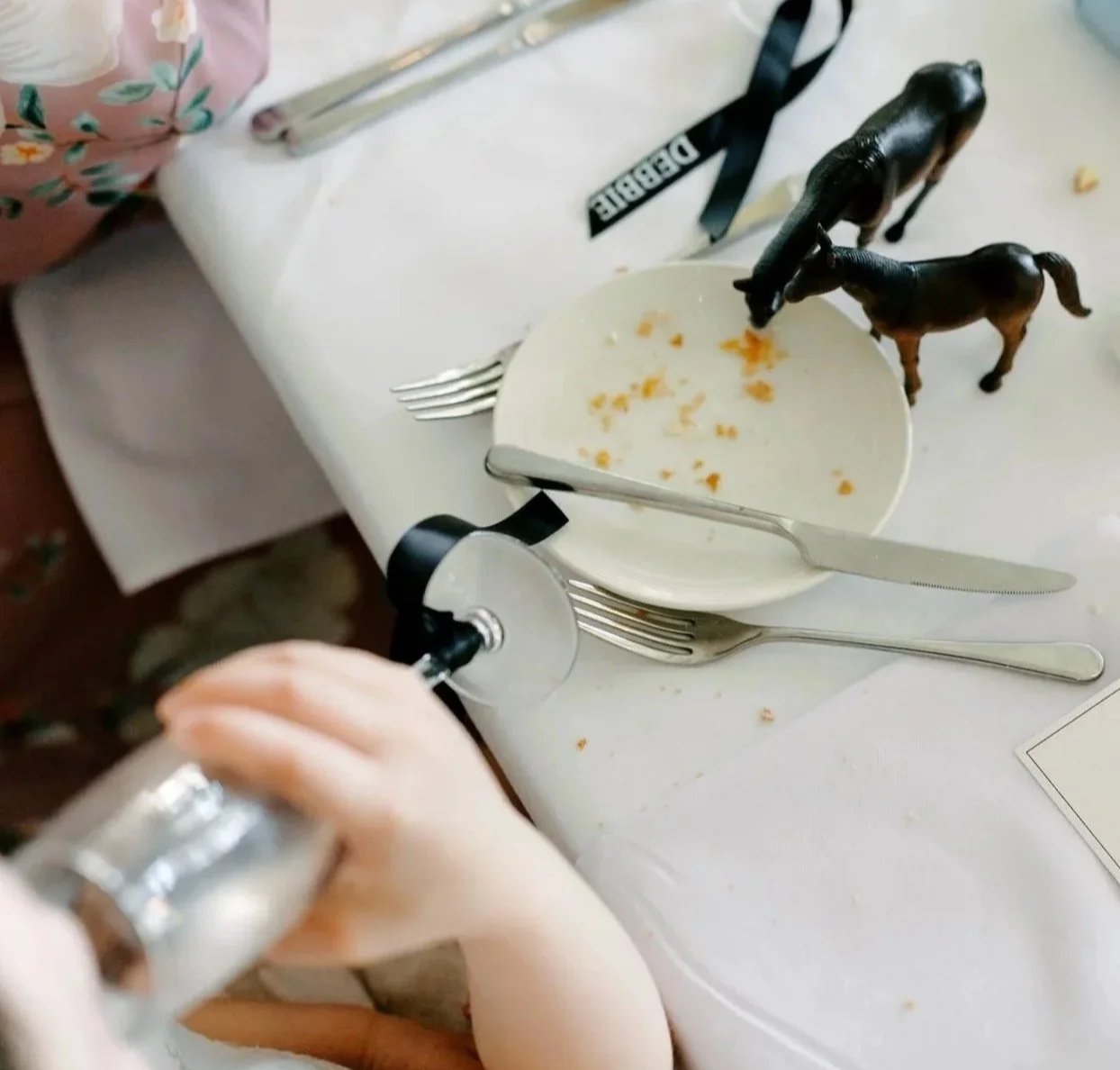 Empty plate with a few crumbs, two forks, and two toy horses on a white tablecloth, with a person holding a glass bottle in the foreground.