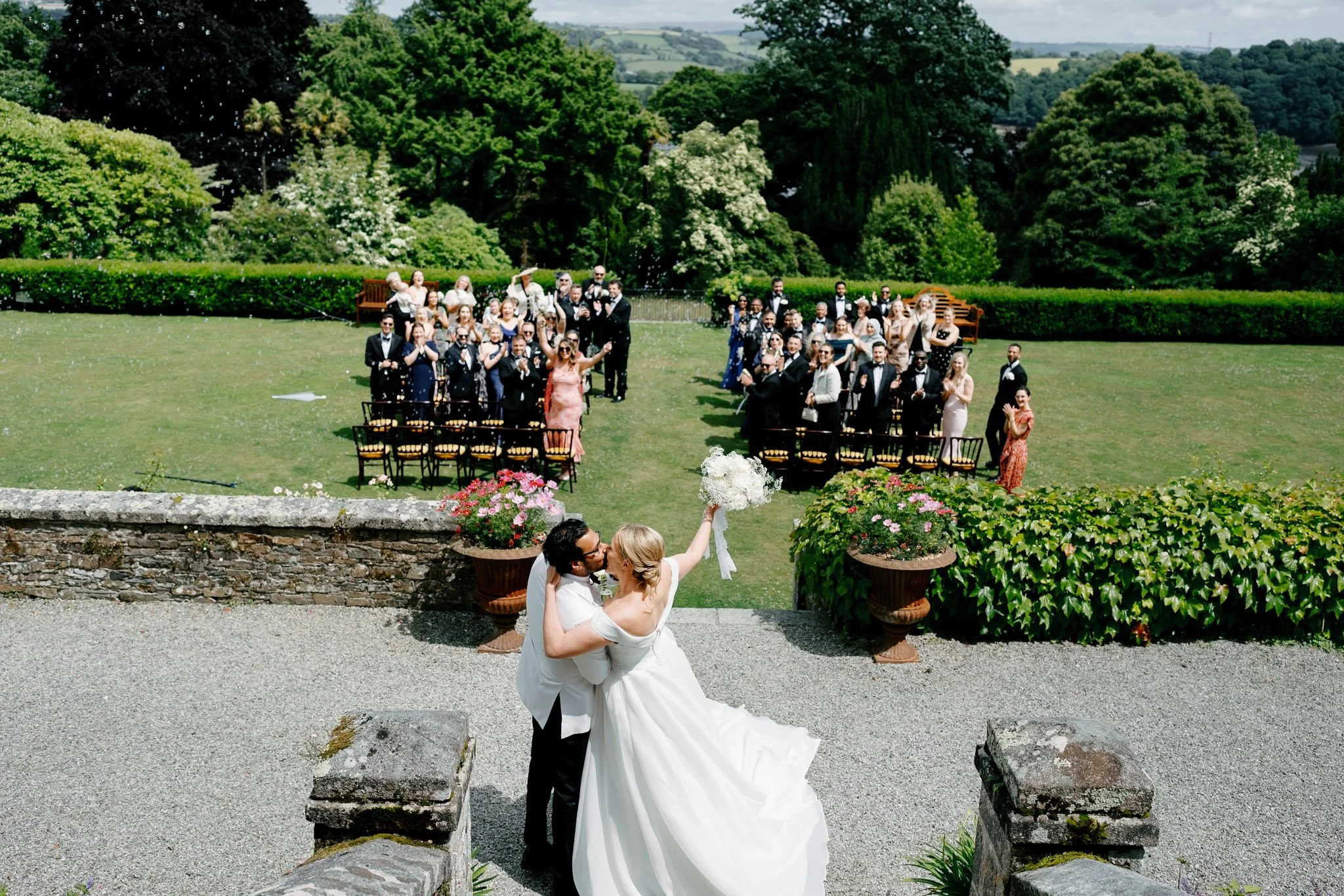 A newlywed couple kissing on a stone staircase, with the bride holding a bouquet of white flowers. They are outside on a sunny day, surrounded by guests seated on chairs in a green garden setting, celebrating their wedding.