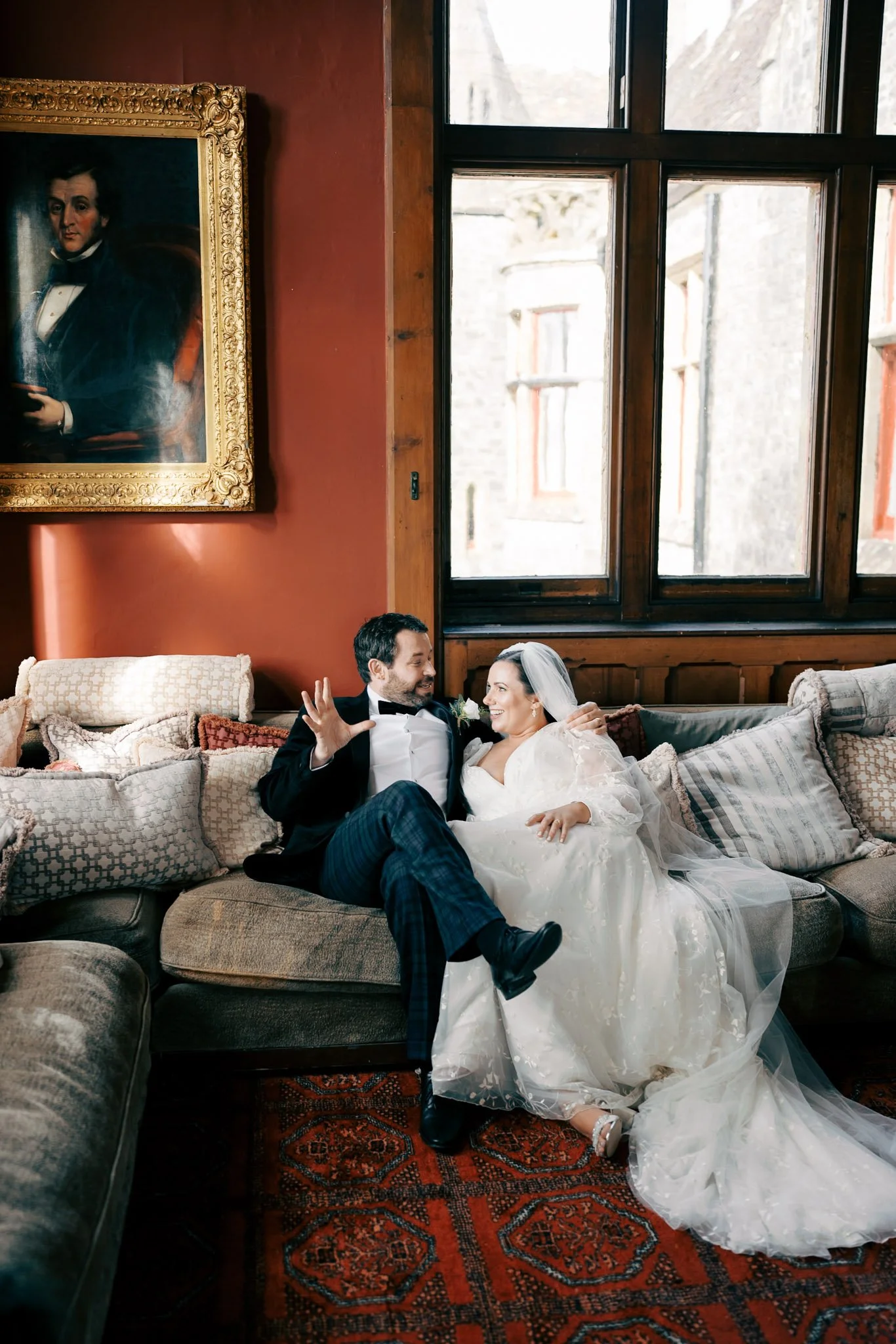 A bride and groom sitting on a vintage sofa in a cozy room with large windows and a portrait painting on the wall. The groom, dressed in a tuxedo, is smiling and gesturing, while the bride, in a white wedding gown, is smiling back at him.