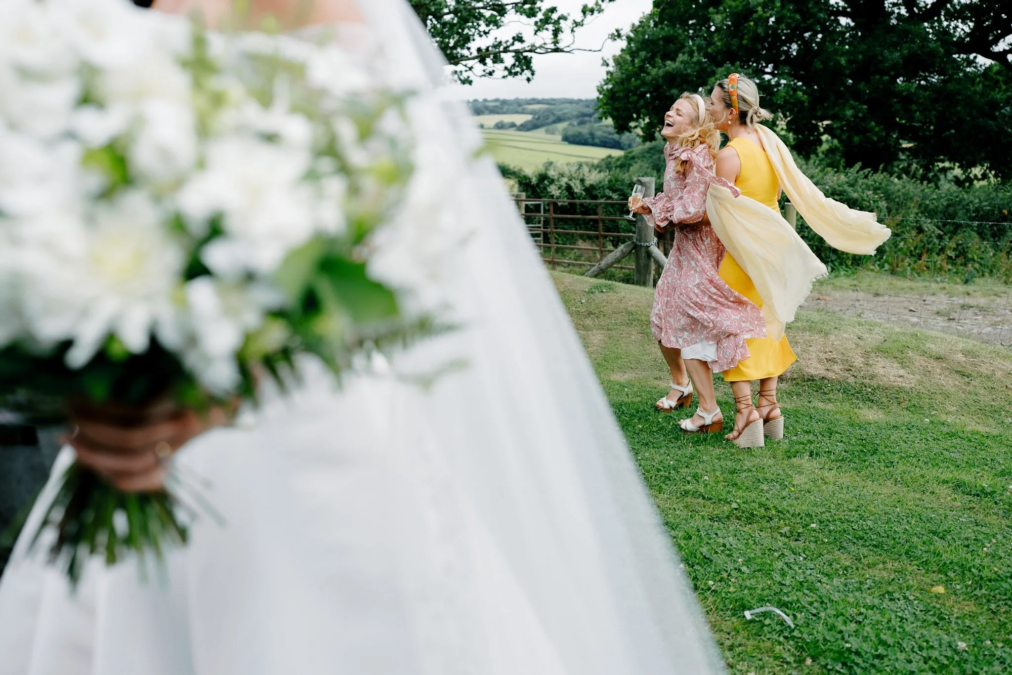 Two women in colorful dresses hugging and laughing outdoors near a wooden fence and green hills, with the edge of a floral wedding arrangement in the foreground.
