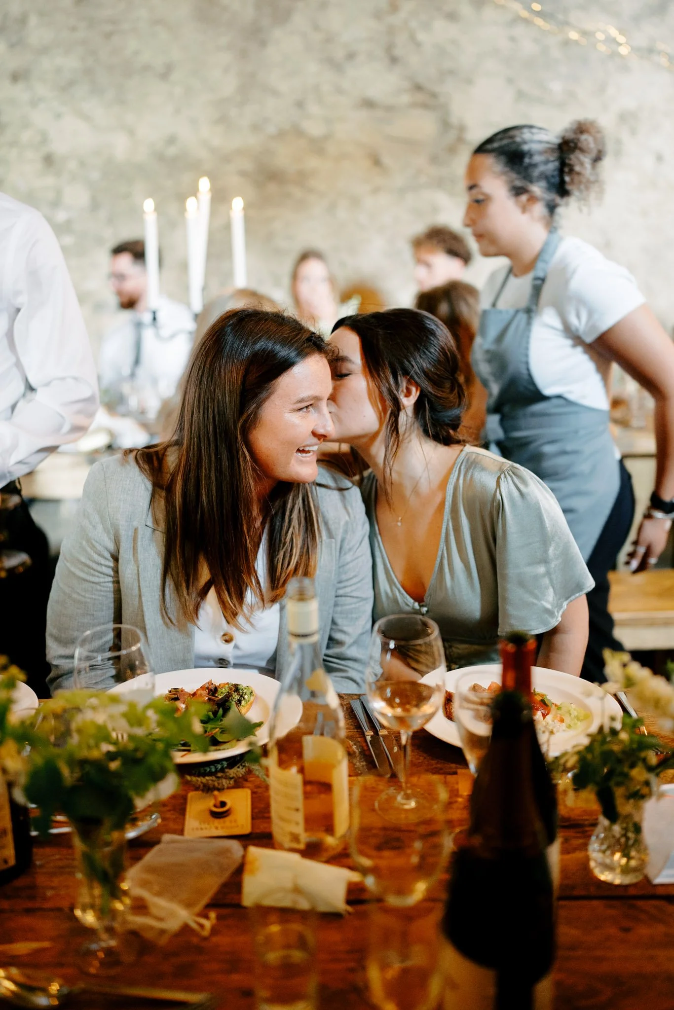 Two women sharing a kiss on the cheek at a dinner table during a wedding celebration in Devon