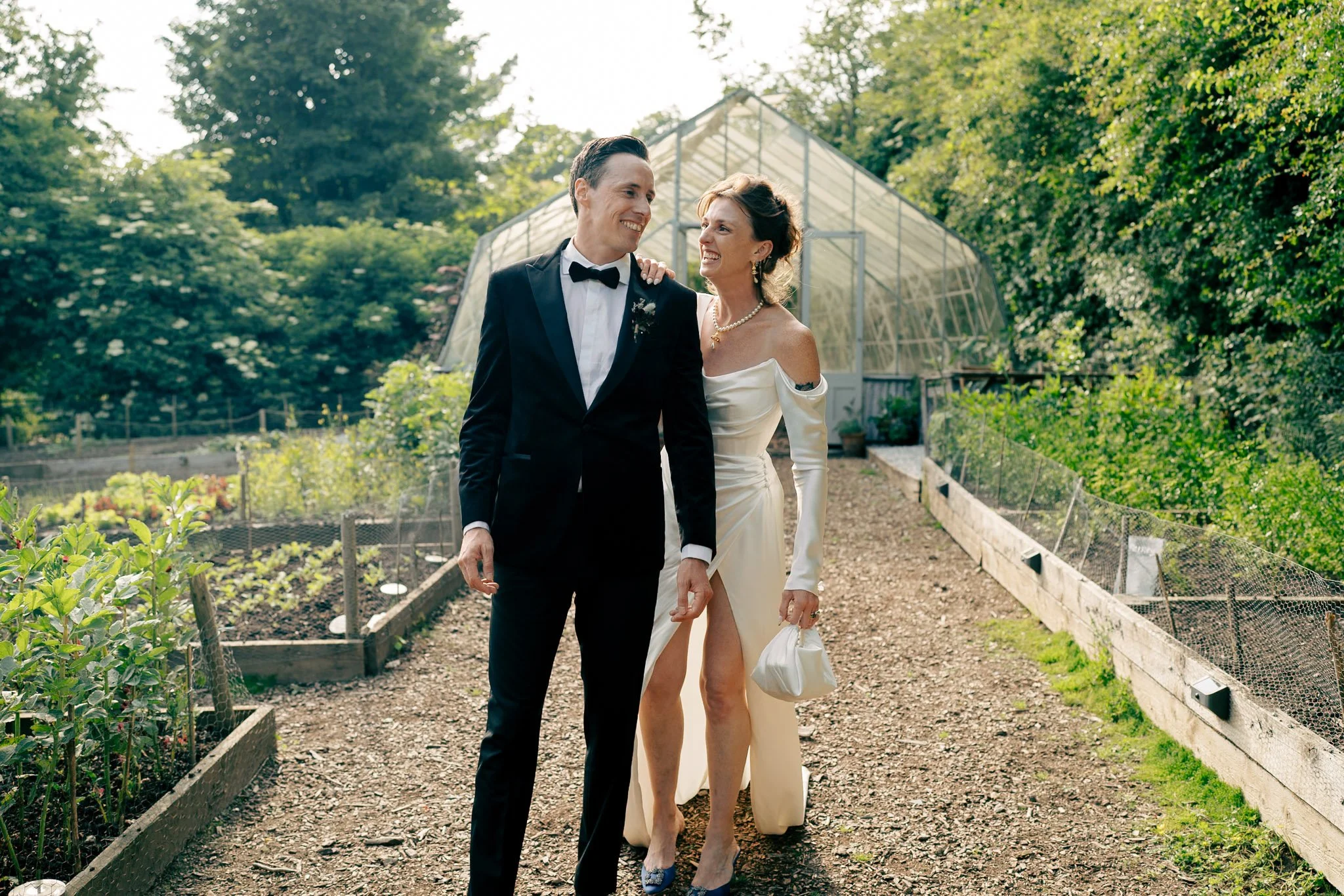 A couple dressed in wedding attire walking on a garden path with greenery and a greenhouse in the background at Nancarrow Wedding venue