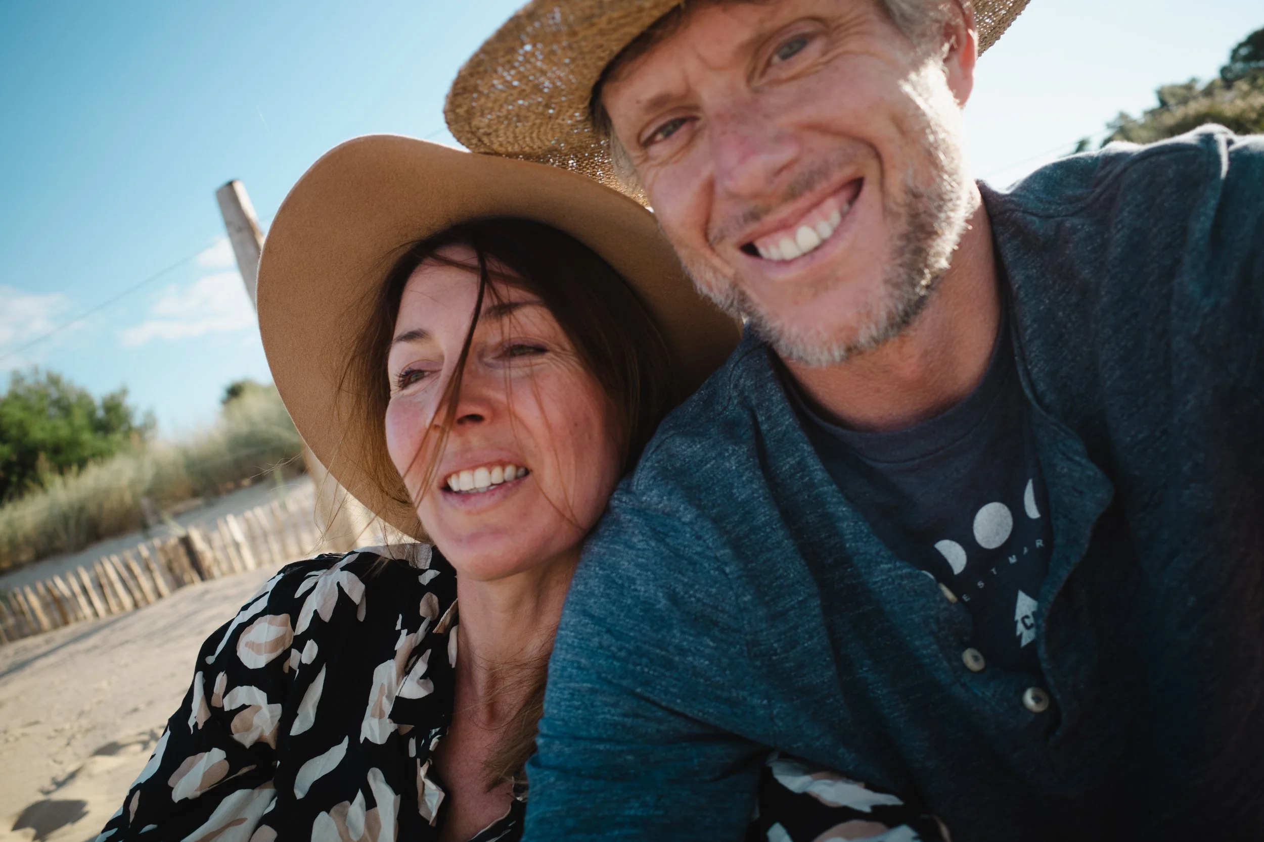 A smiling man and woman taking a selfie together outdoors on a sunny day, with the man wearing a hat and the woman wearing a wide-brimmed hat.