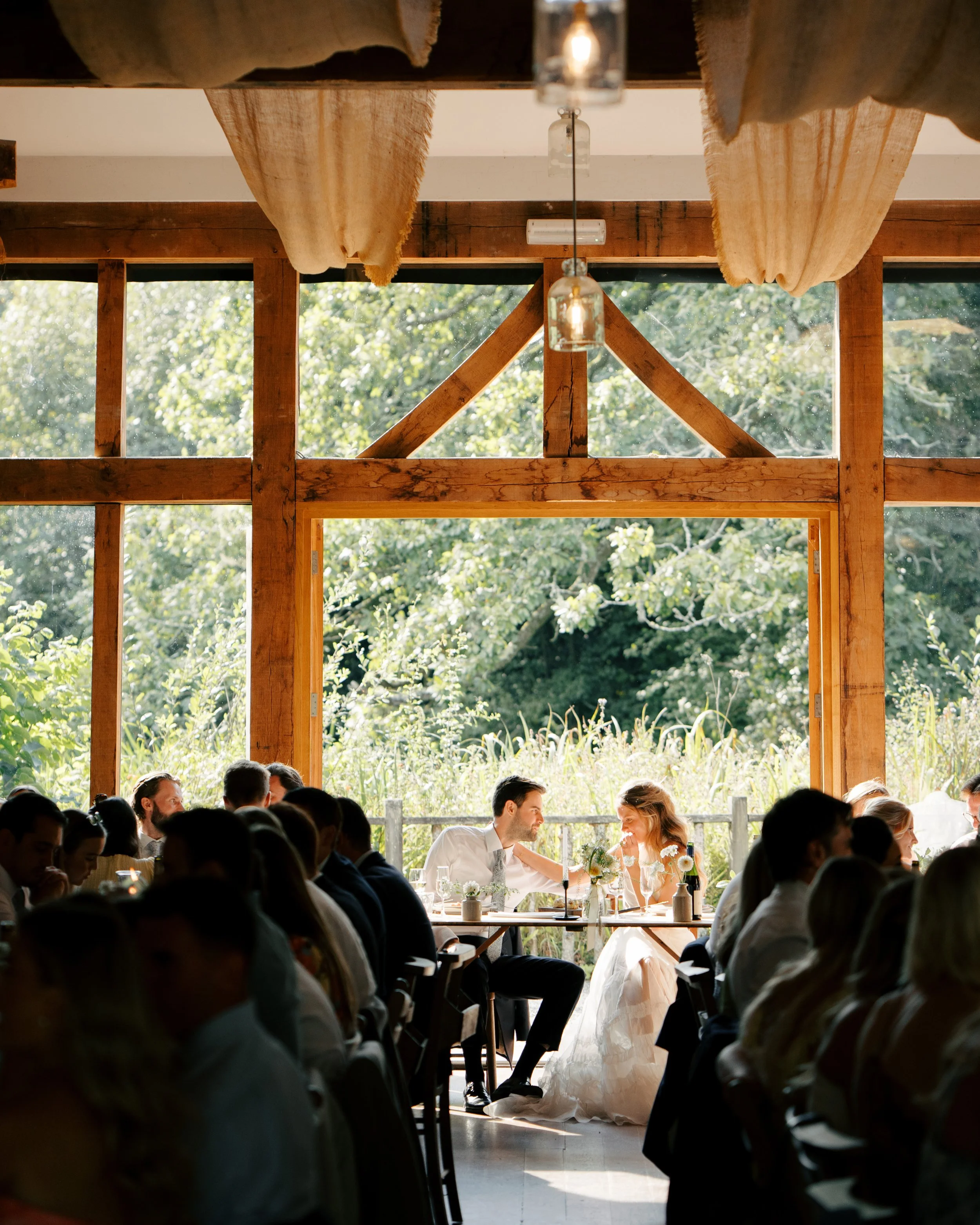 Bride and groom sitting at a table during their wedding reception in a rustic venue with large windows and greenery outside, surrounded by seated guests.