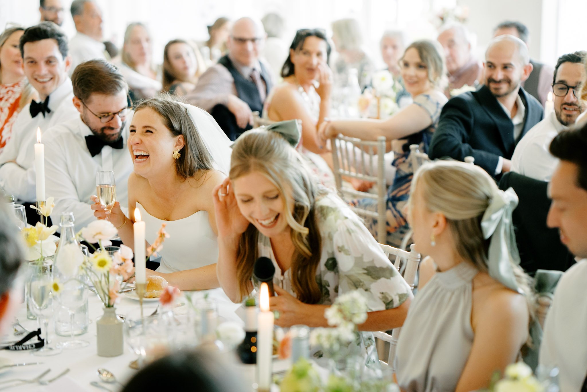 Guests at a wedding reception, including a woman in a wedding dress laughing and a woman speaking into a microphone, seated at a table with floral arrangements and candles.