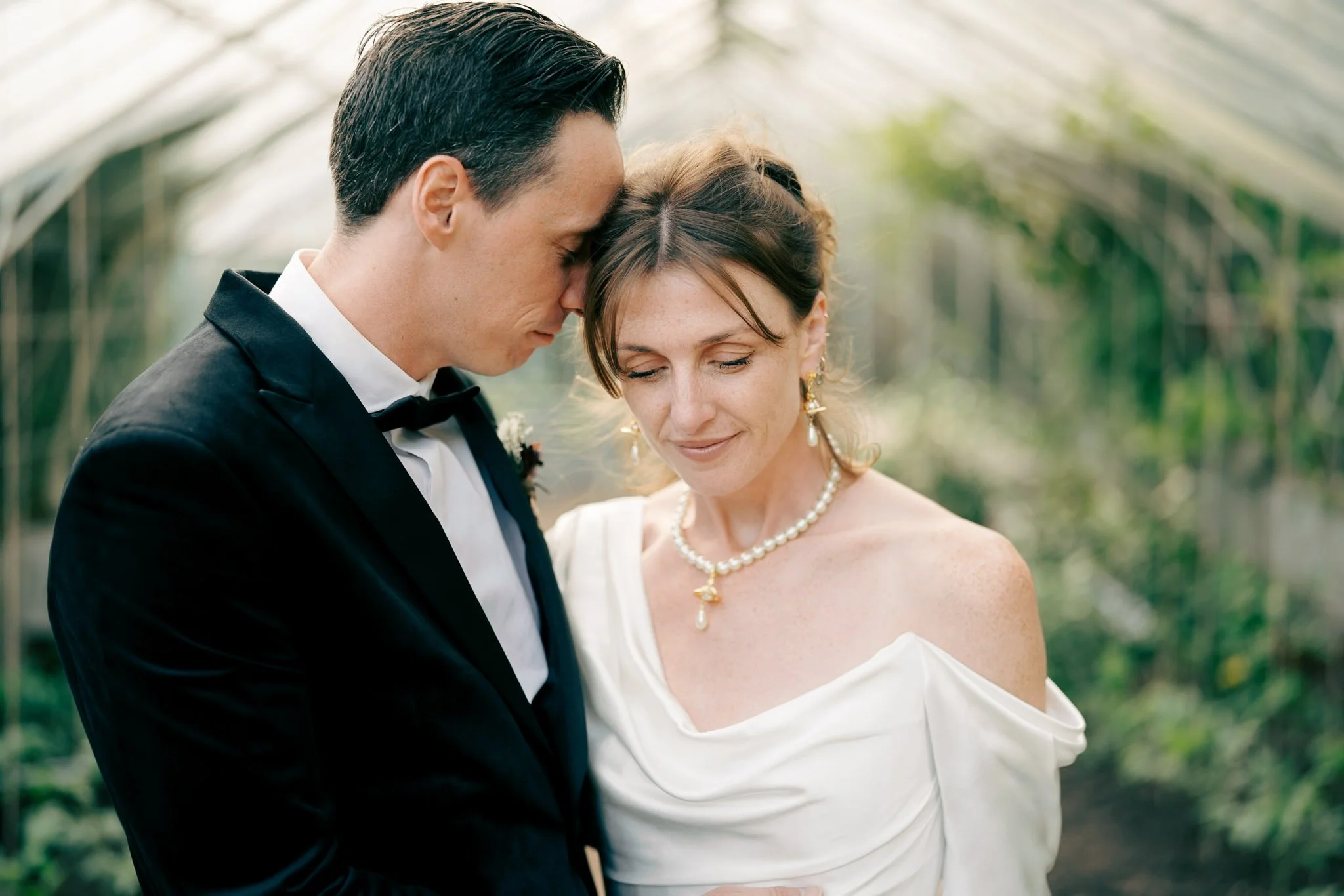 A bride and groom share a tender moment with their foreheads touching in a greenhouse, both with eyes closed, dressed in wedding attire.