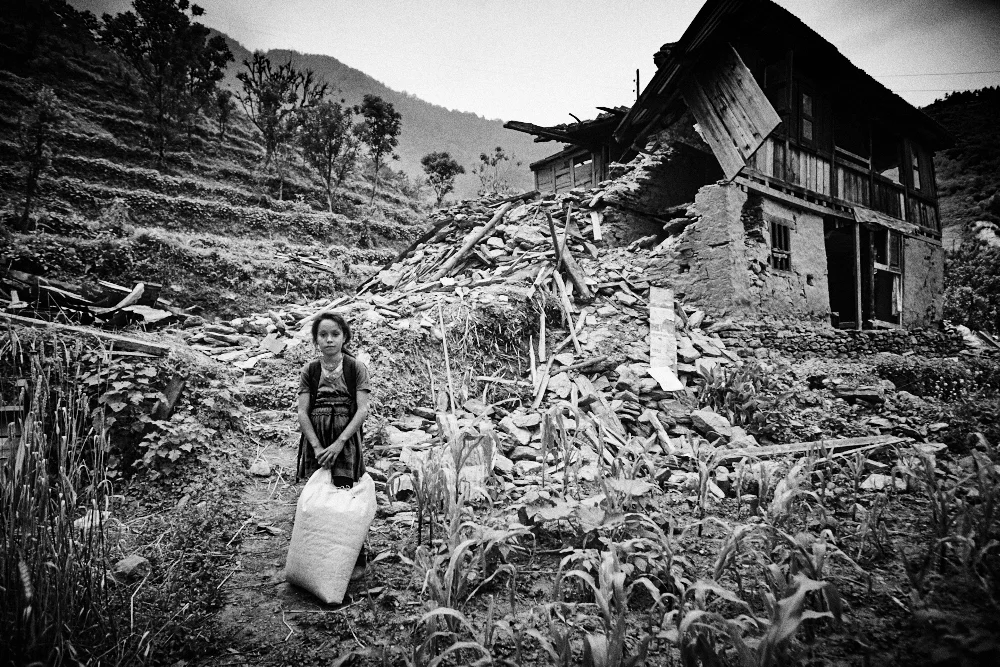Menuka stands with a bag of rice. Behind her stands what remains of her house in Fulping Katti.