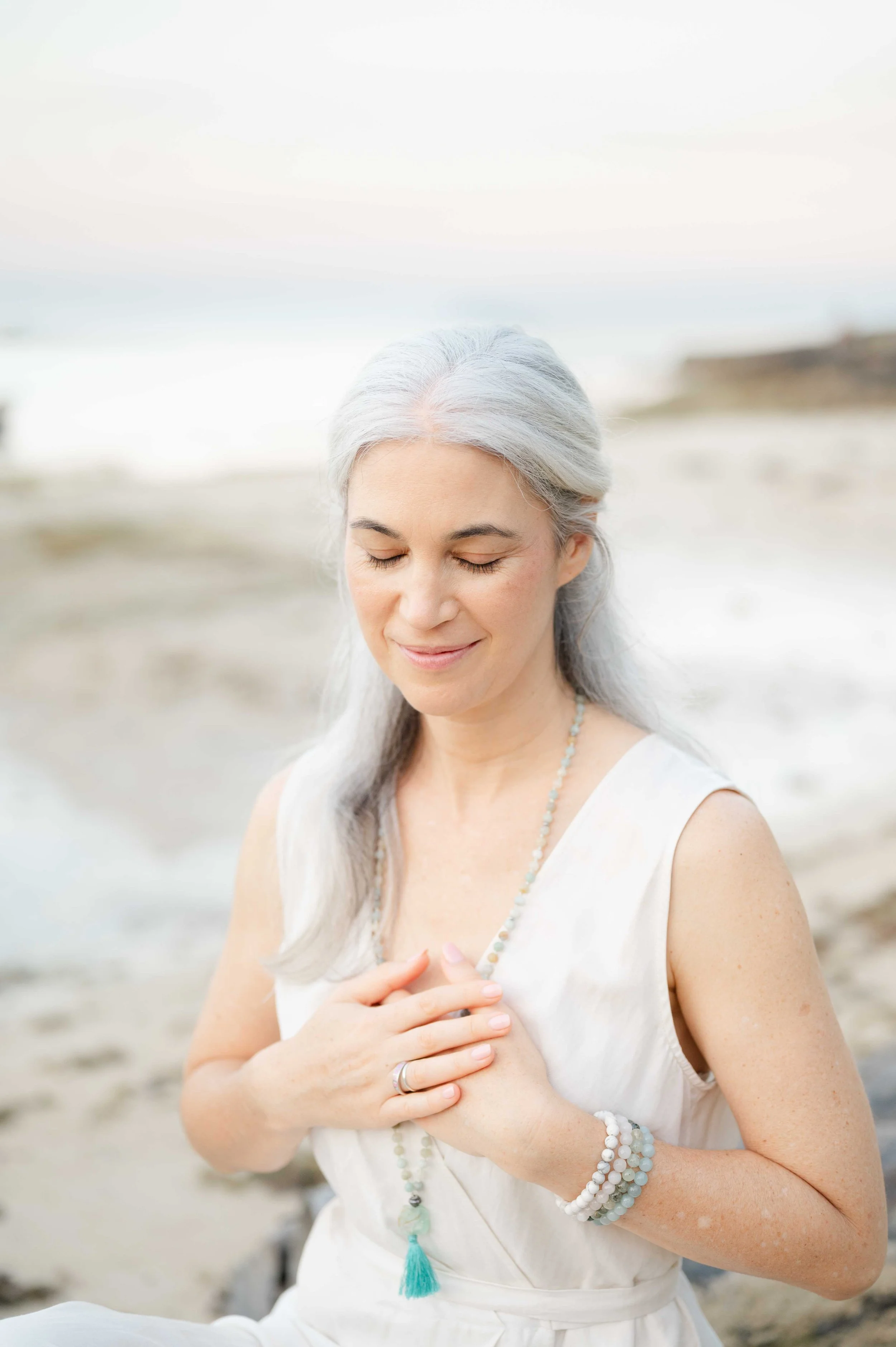 Woman with white hair looking down with soft smile, hands on her heart