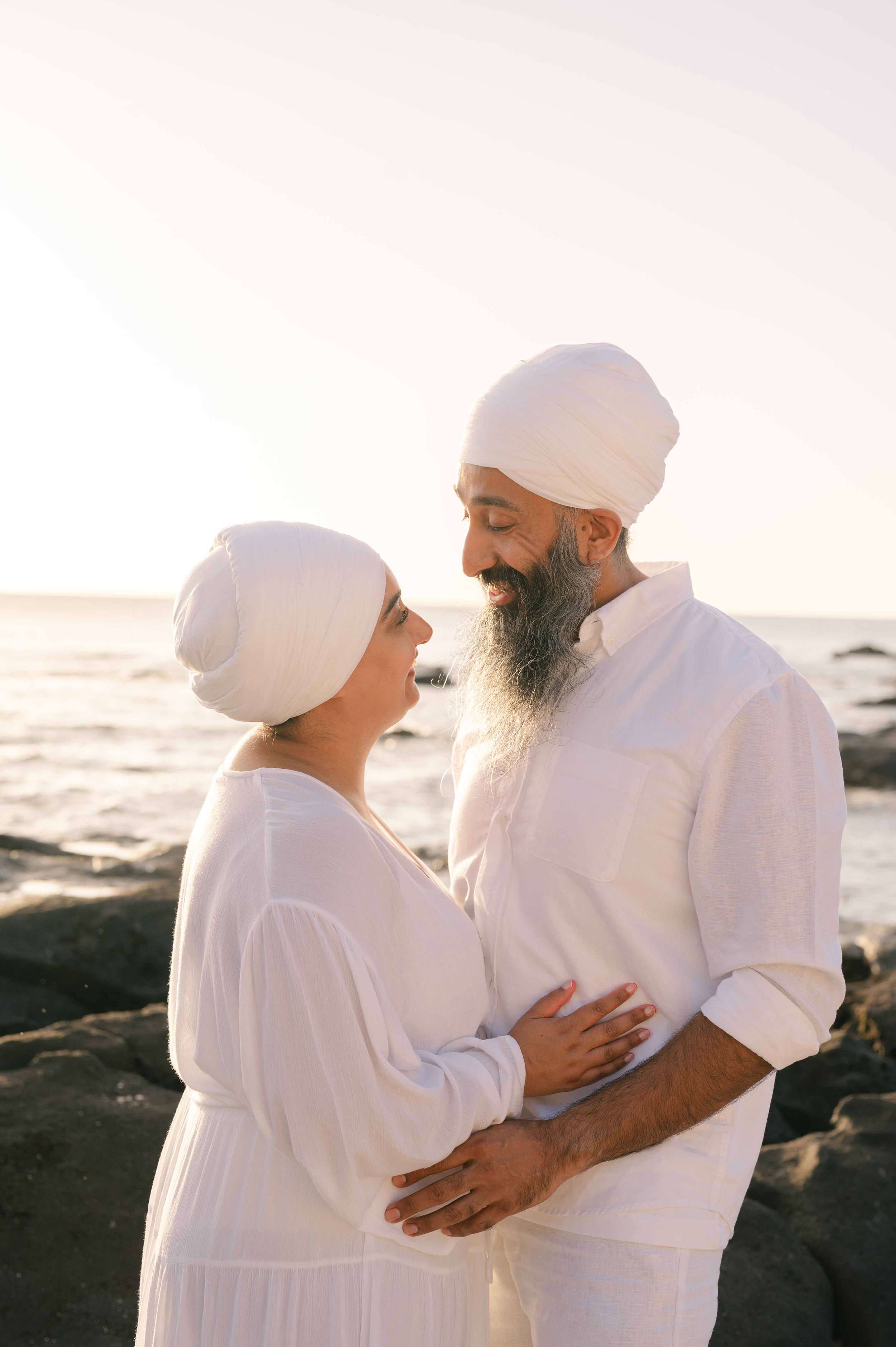 A couple wearing white, standing close on a beach at sunset, gazing into each other's eyes.