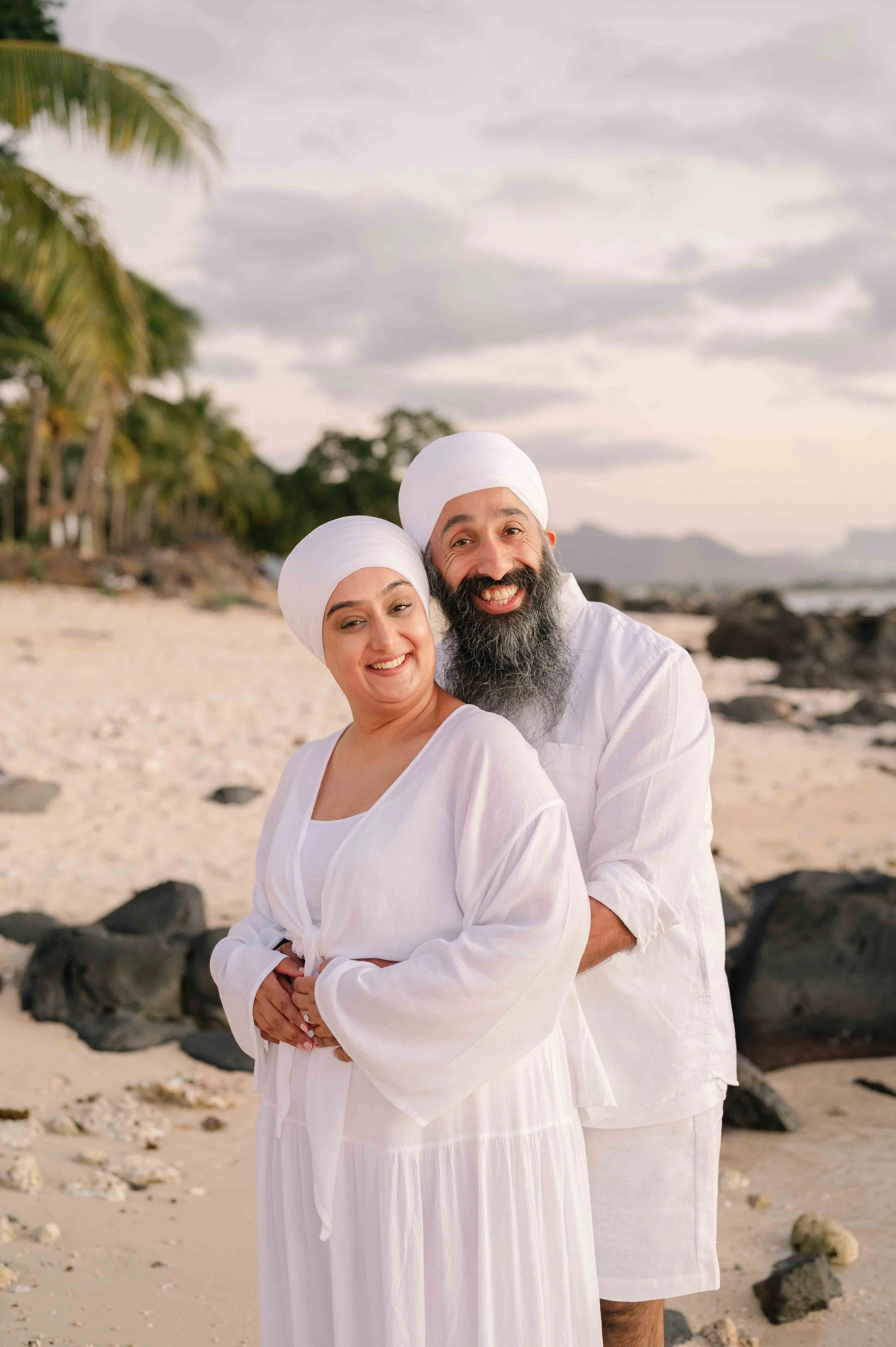 A smiling couple wearing white clothing and turbans on a beach with palm trees and rocks, during sunset or sunrise.