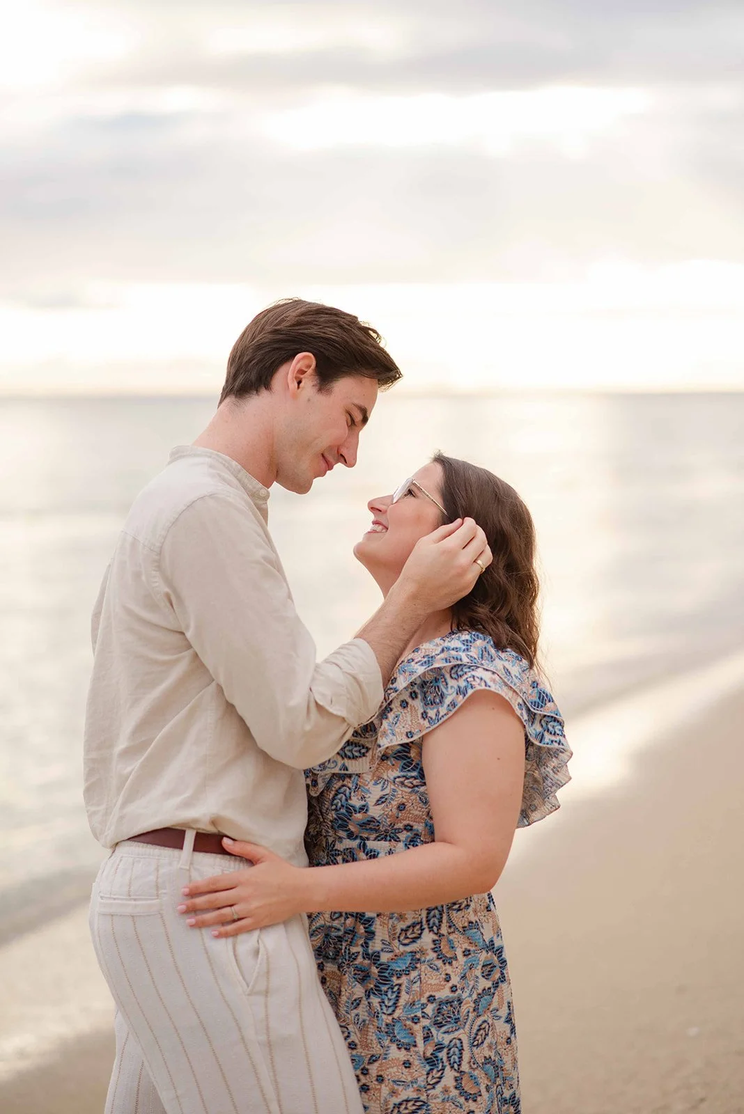 A man and woman on a beach, looking at each other and smiling, with the ocean and a cloudy sky in the background.