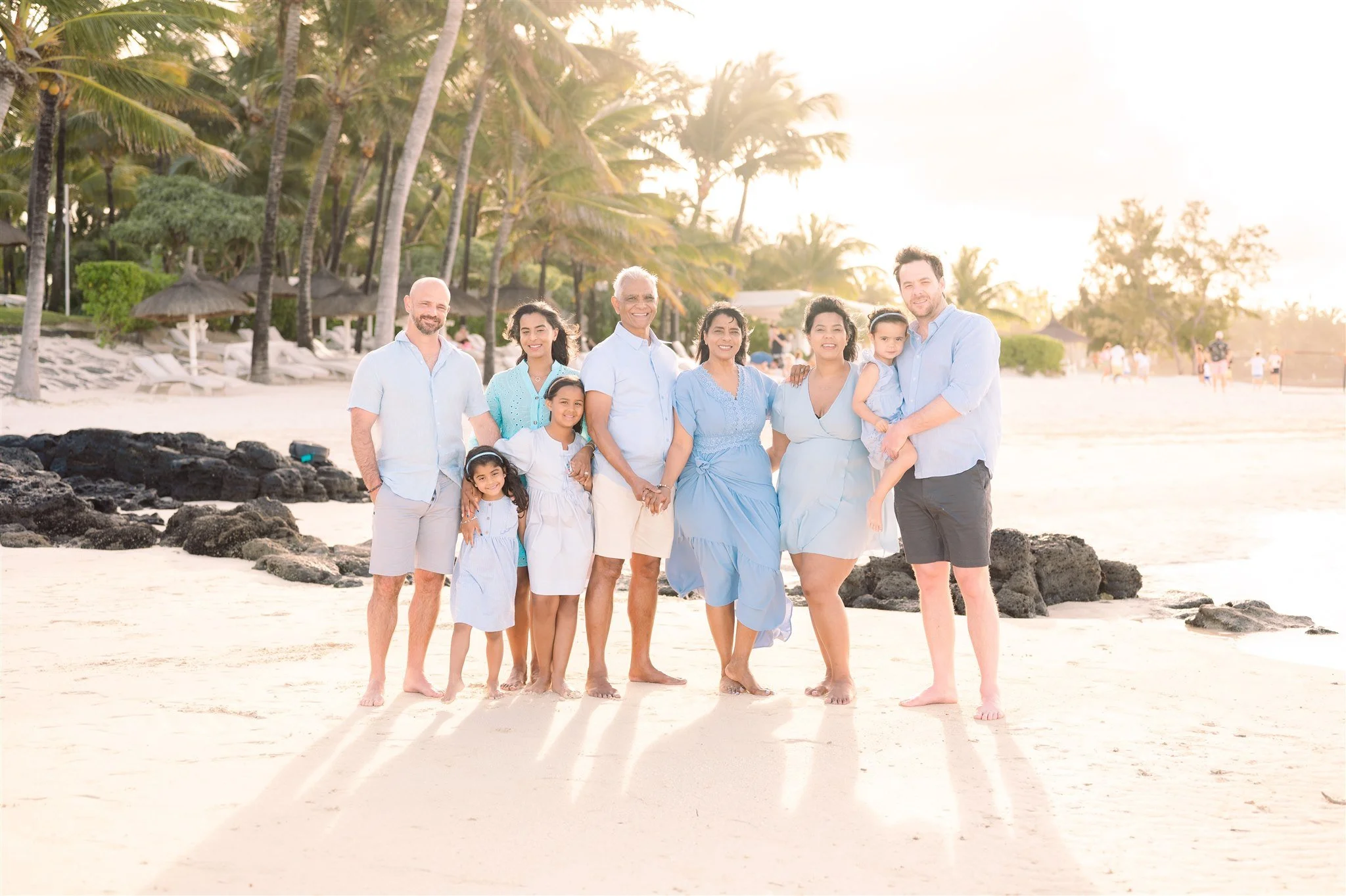 Family gathering on a beach during sunset, with palm trees and rocks in the background, all dressed in light blue and white clothing.