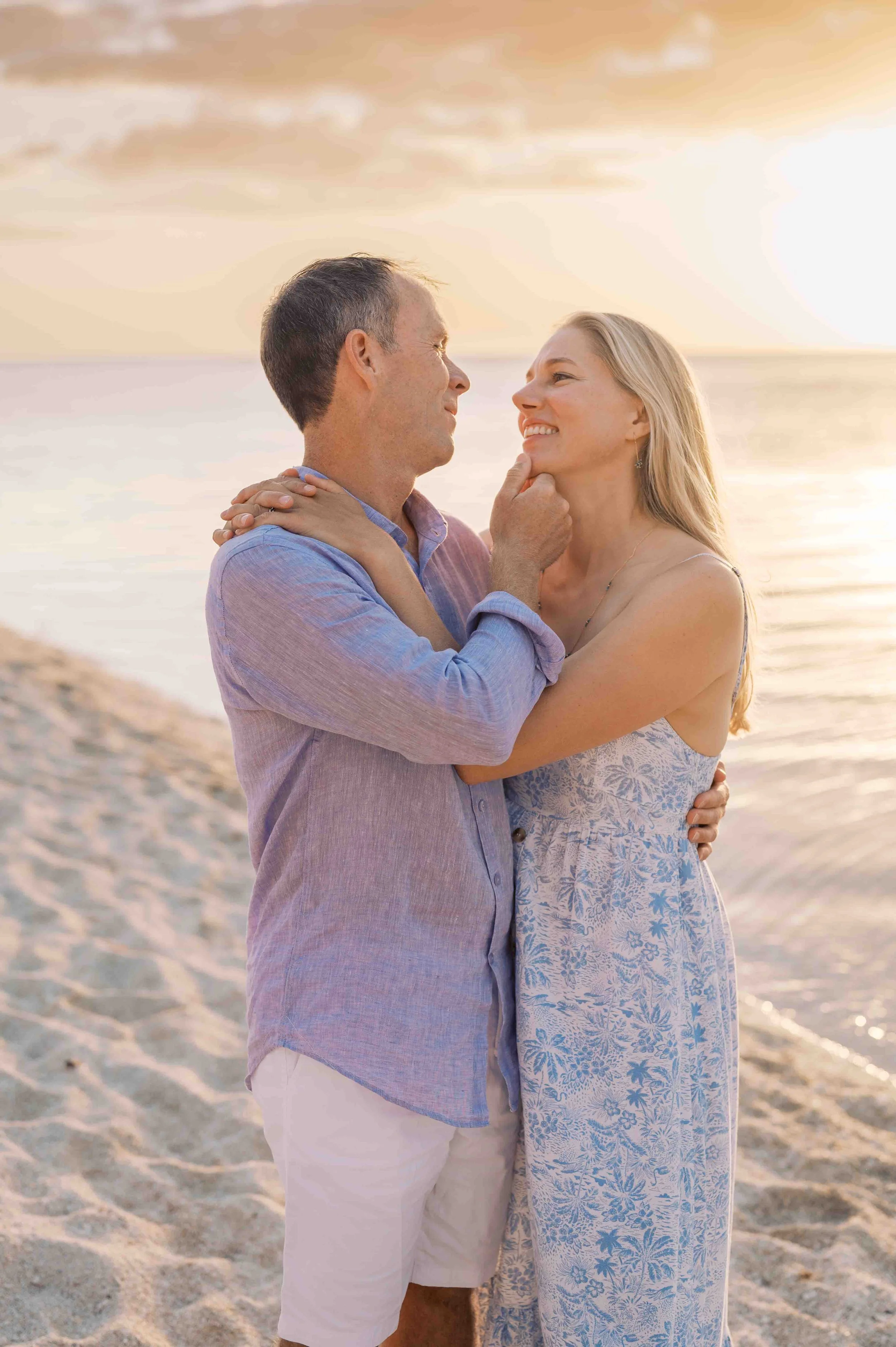 A couple embracing on the beach at sunset, with a man holding the woman's chin and smiling while she also smiles, and the ocean and sky in the background.