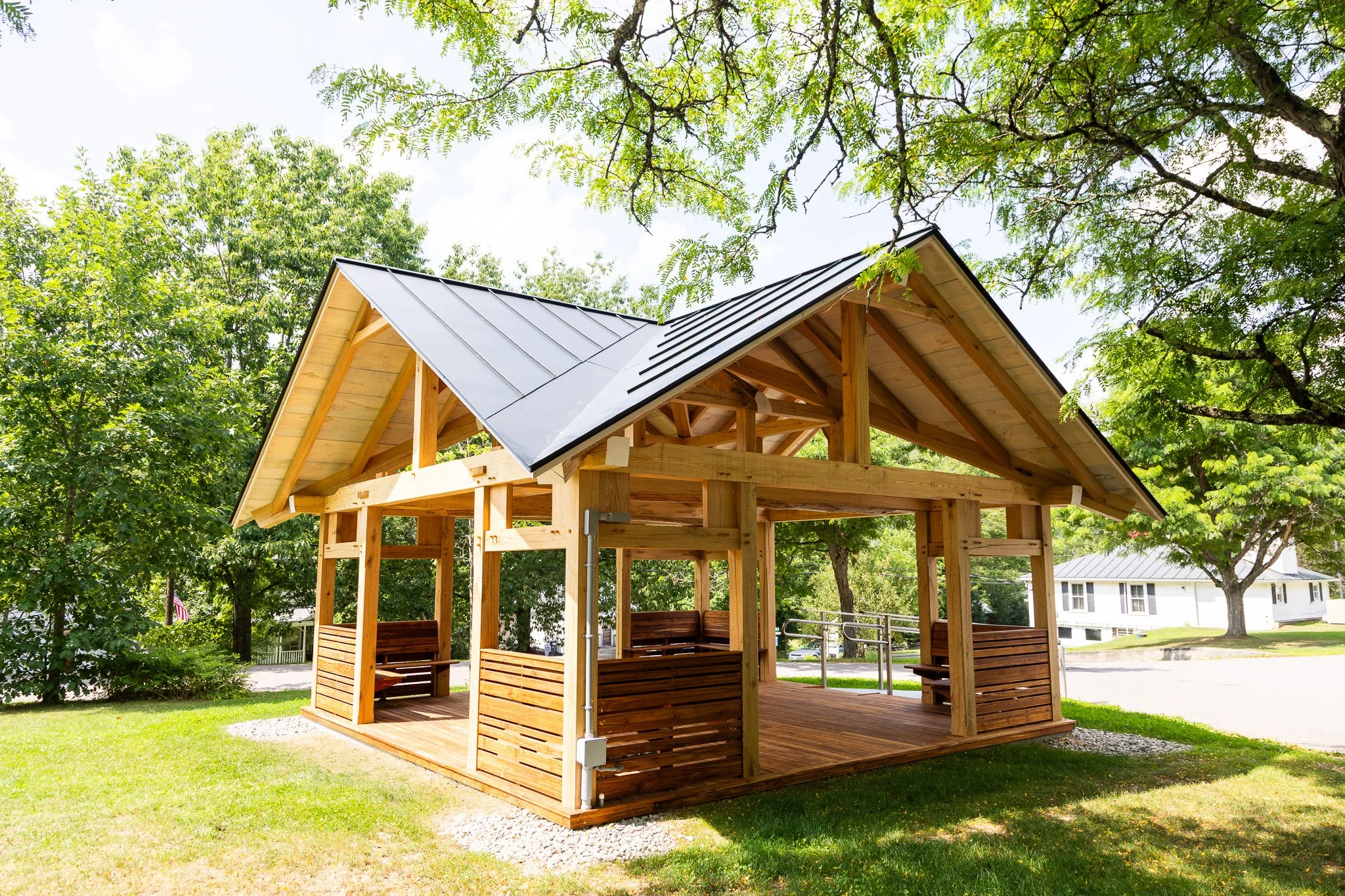 A small wooden pavilion with a metal roof, surrounded by green trees and grass. Designed by Corrigan & Klein Artisan Builders.
