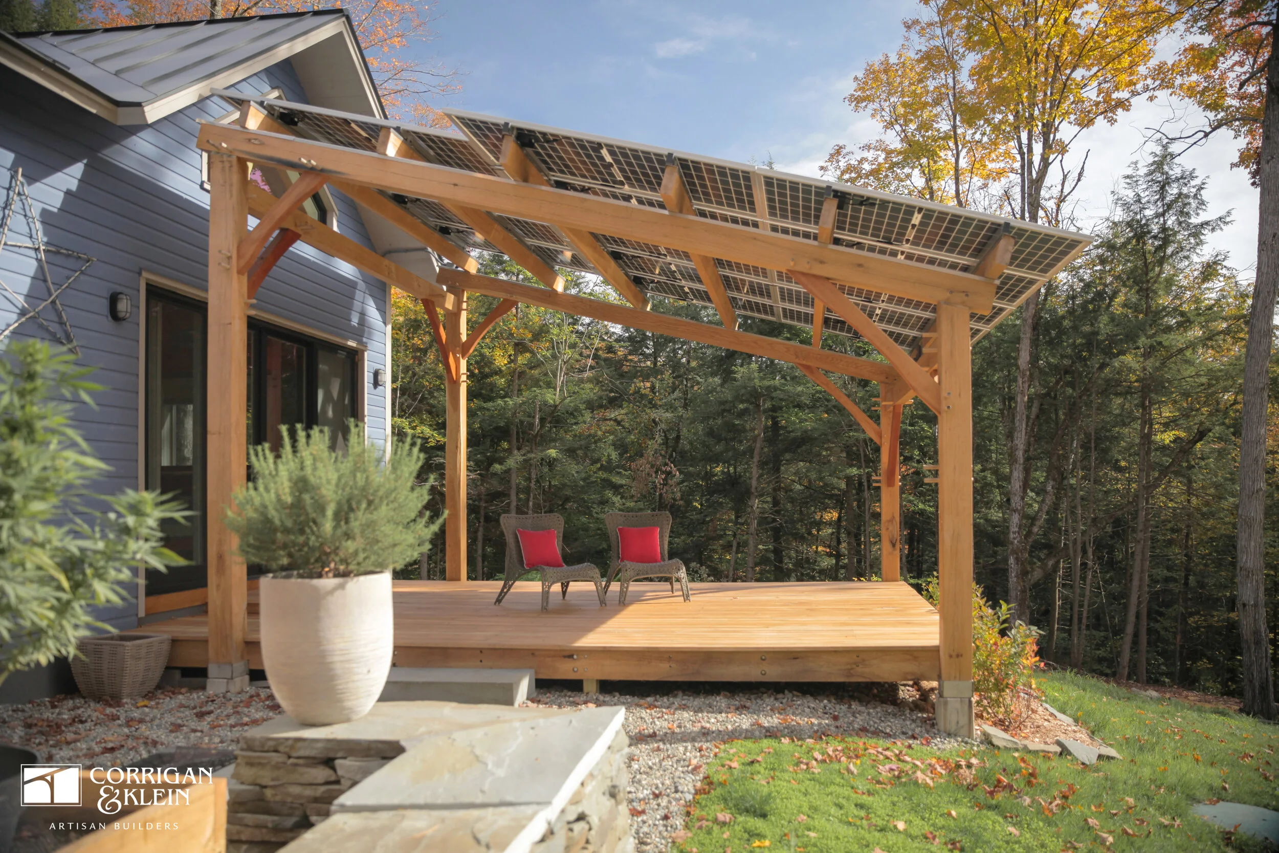 A wooden deck under a pergola with solar panels, surrounded by trees with autumn foliage. Designed by Corrigan & Klein Artisan Builders.