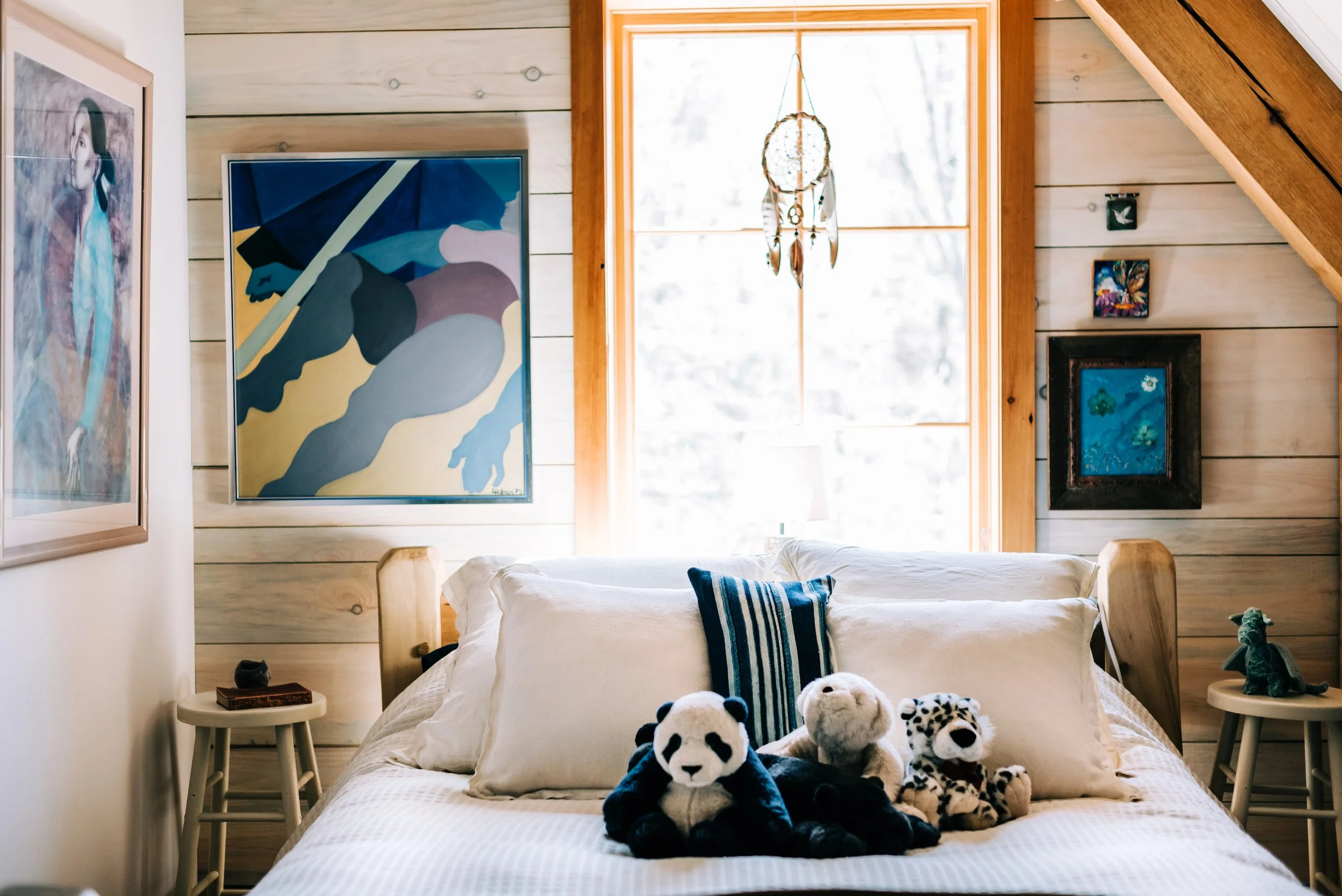A cozy bedroom corner with a bed shown from the foot and two small side tables flank the bed. The wall behind the bed is made of light wood paneling. A large window with a wooden frame lets in natural light. Designed by Corrigan & Klein Artisan Build