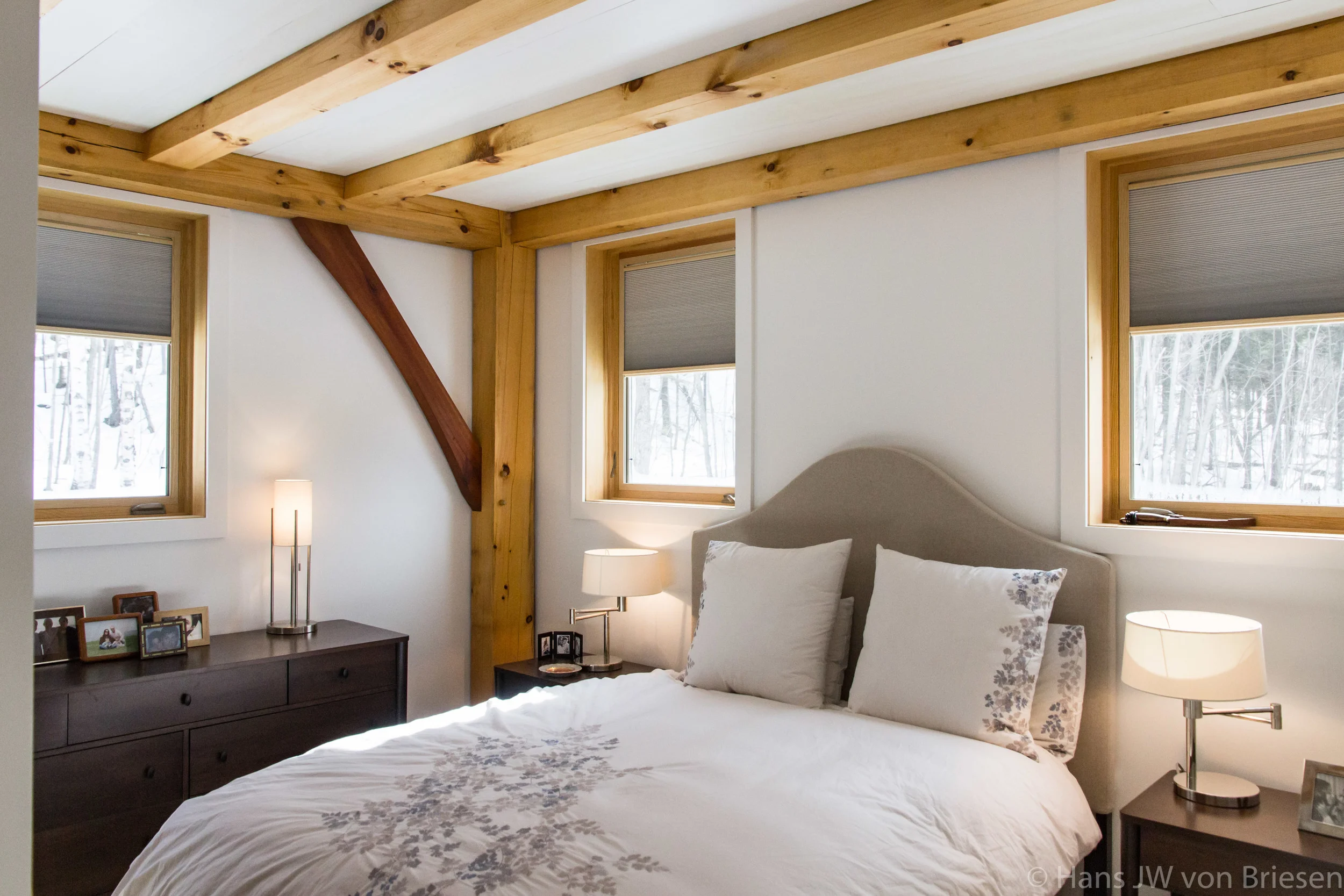 A cozy bedroom with a bed, and a beige headboard. Wooden beams and window frames add rustic charm. Nightstands with lamps and personal photos are beside the bed. Designed by Corrigan & Klein Artisan Builders.