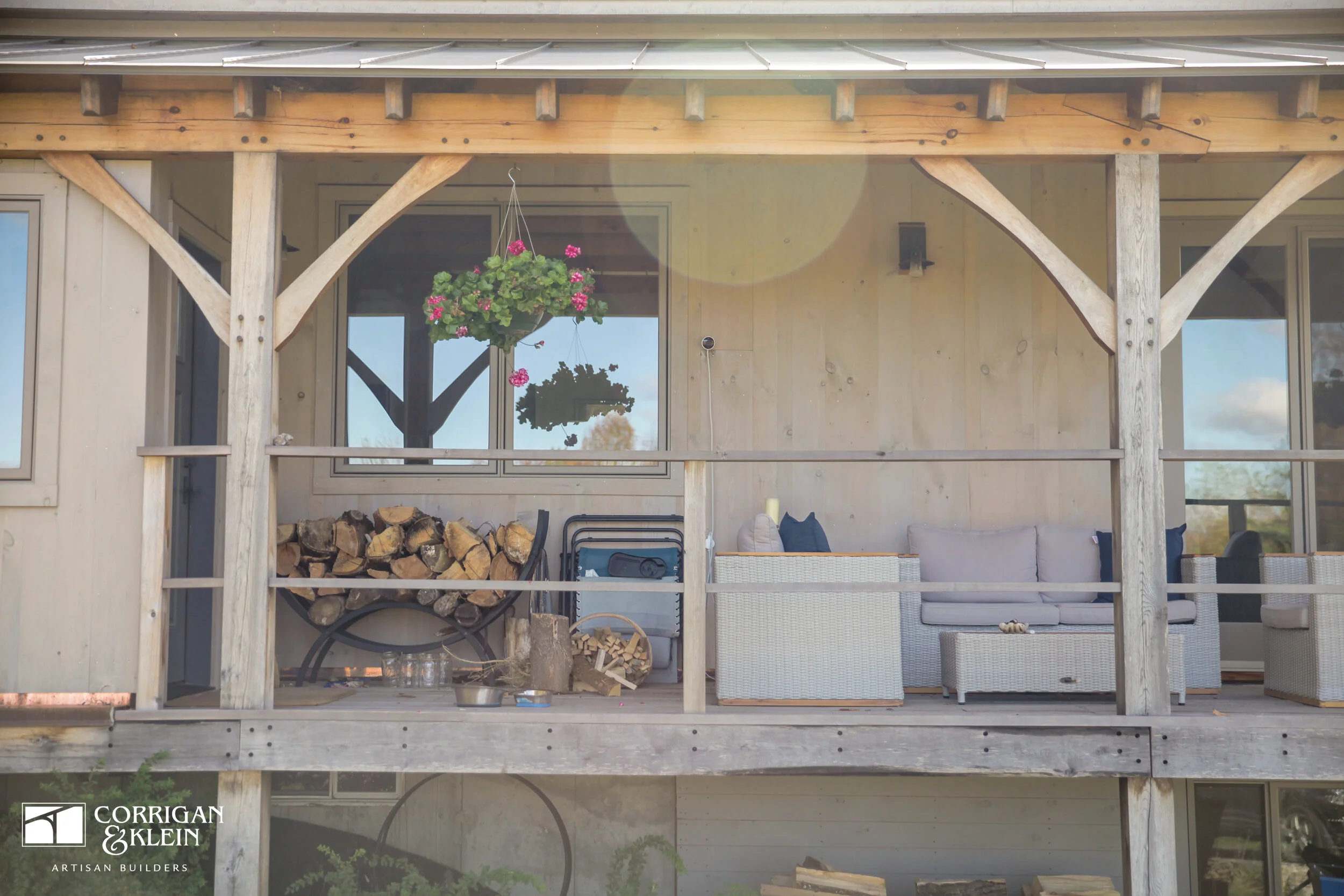 Front porch of a wooden house with outdoor furniture, a hanging plant, and stacked firewood, under a metal roof. Designed by Corrigan & Klein Artisan Builders.
